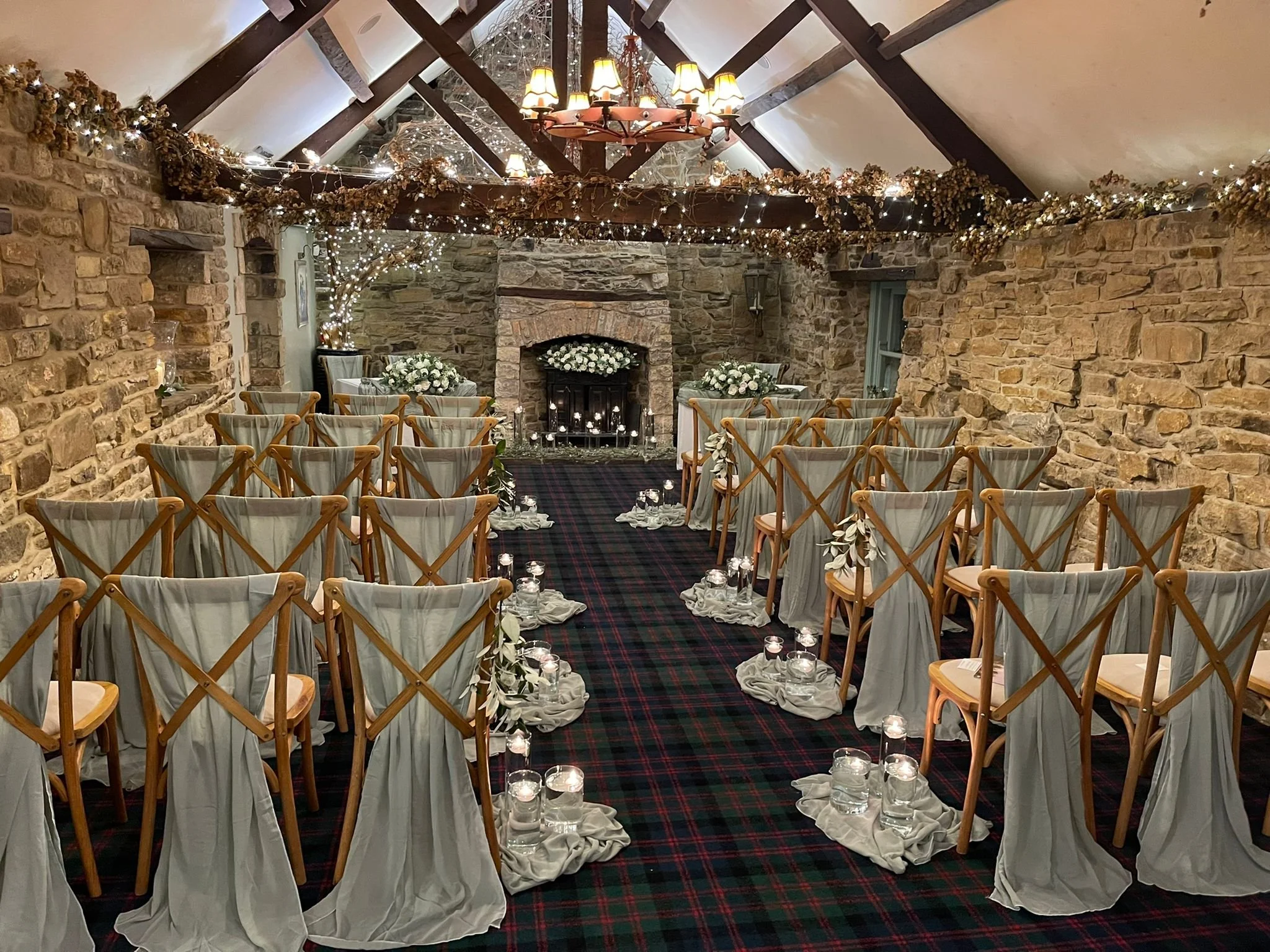 Indoor wedding ceremony setup with rows of wooden chairs draped in white fabric, lining a central aisle decorated with candles in jars. The rustic stone walls and ceiling beams are adorned with fairy lights and dried floral garlands, and a cozy stone