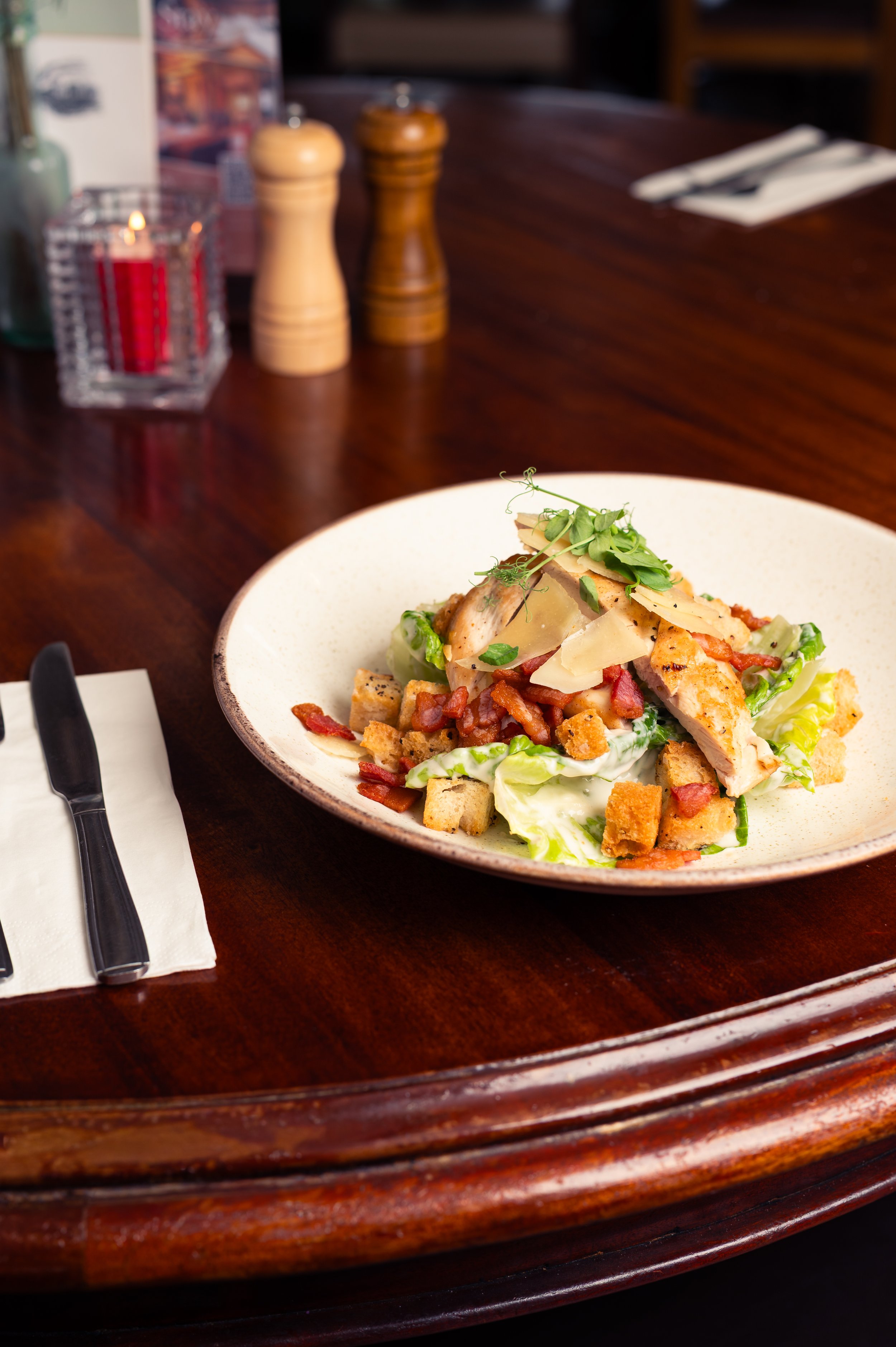 A plate of salad with grilled chicken, bacon, cheese, croutons, and greens on a wooden table with salt and pepper shakers, a candle, and napkins.