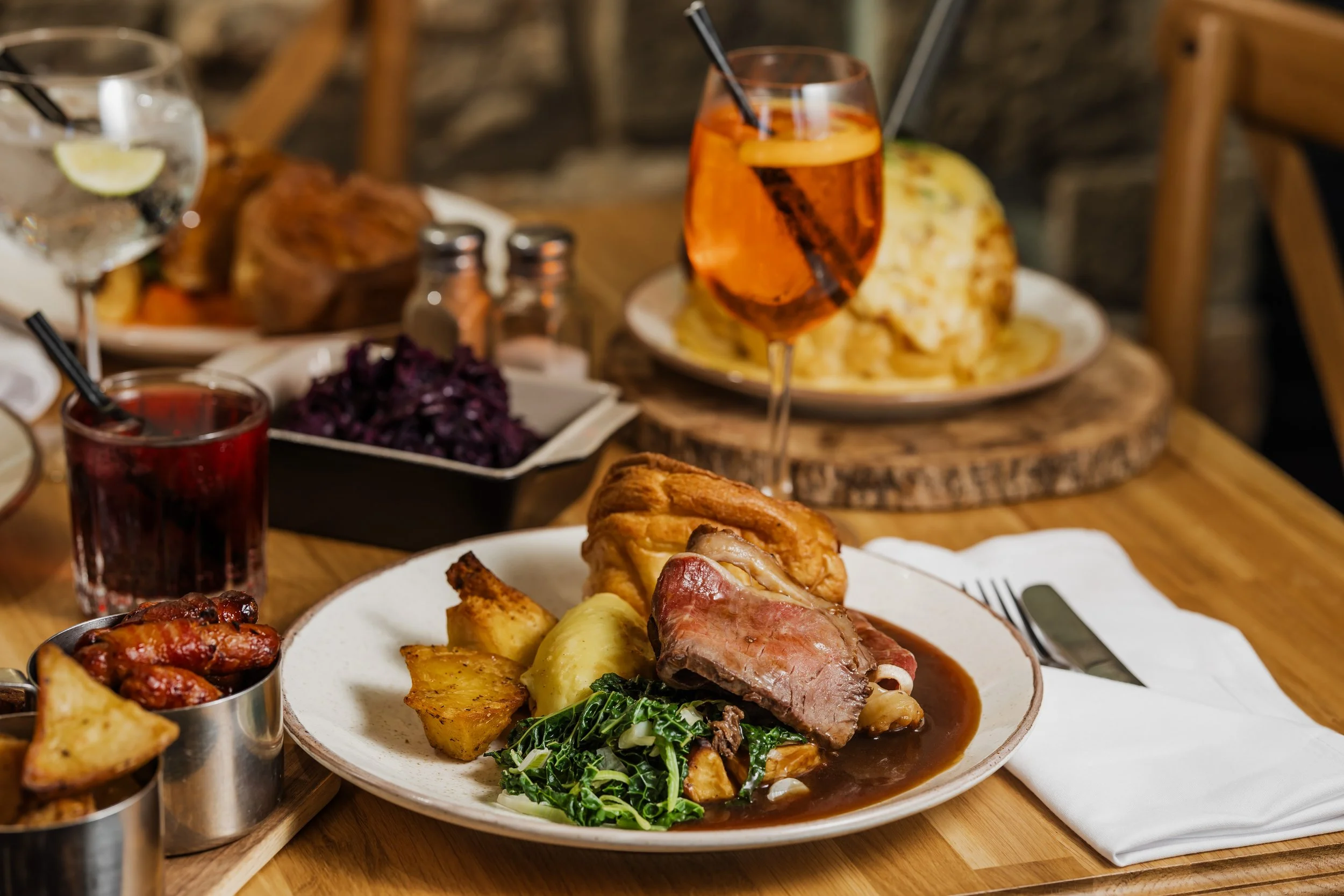 A dinner table with roast beef, mashed potatoes, roasted potatoes, sautéed greens, and gravy, along with drinks including a glass of wine and a dark beverage, with various side dishes and condiments.