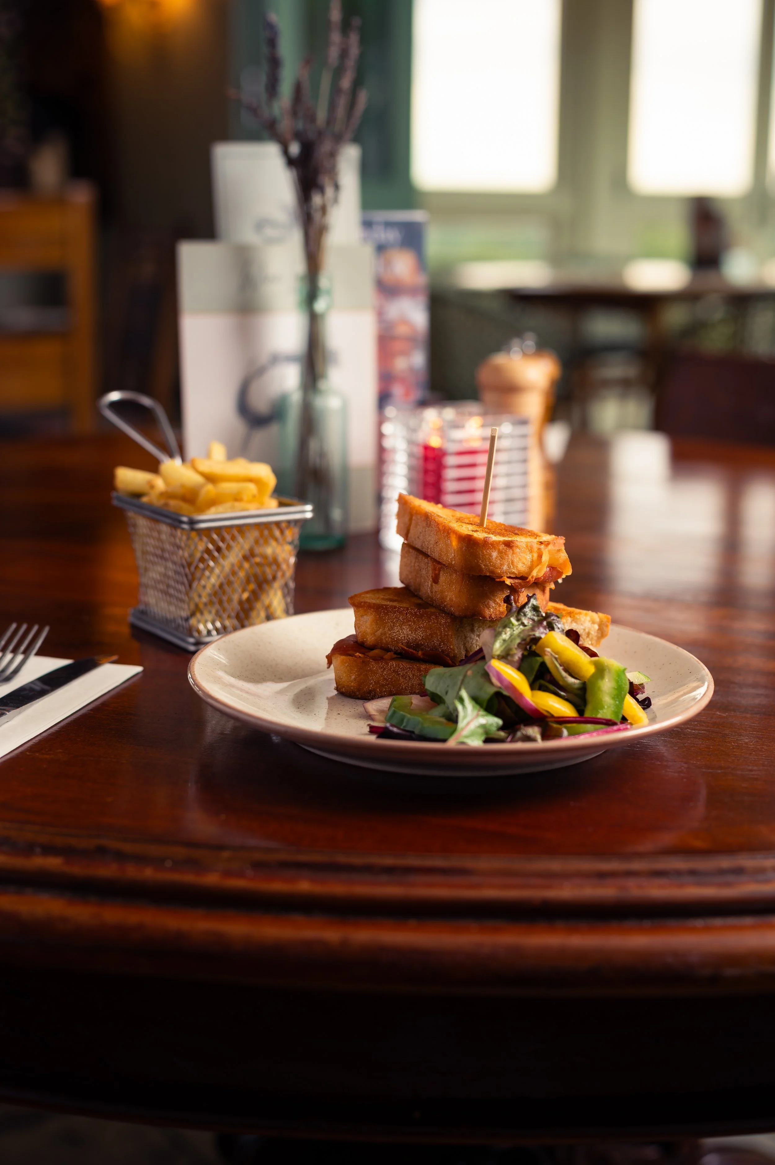 A plate of club sandwich with lettuce, tomato, and pickles, served with a side salad. In the background, there is a basket of French fries, a bottle of water, and a blurred view of a café interior with large windows.