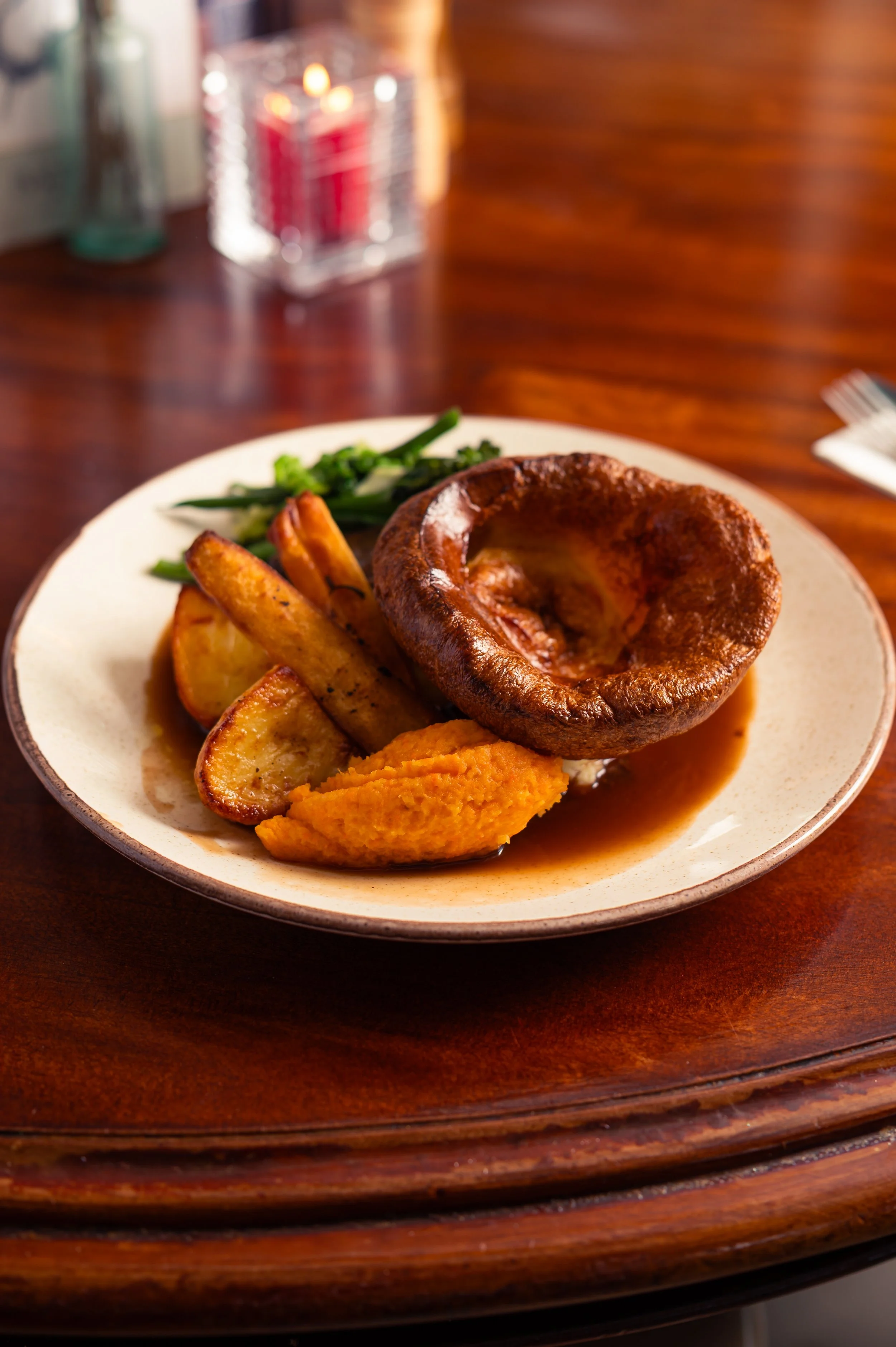 A plate of food with roasted potatoes, green beans, a large Yorkshire pudding, and a piece of fried fish, set on a wooden table.