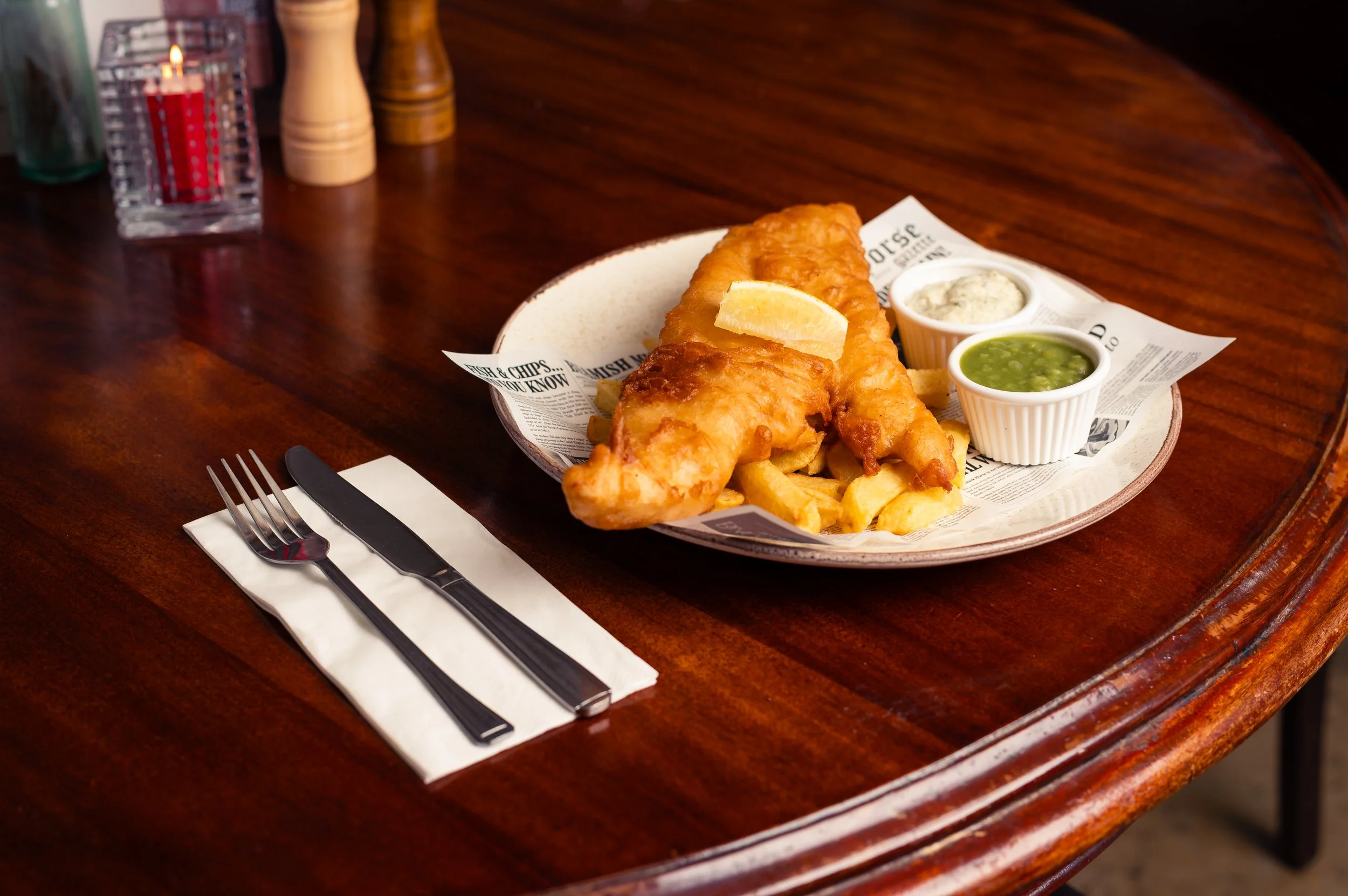 Plate of fish and chips with lemon wedge and three dipping sauces, on a wooden table with salt and pepper shakers and water glasses in the background.