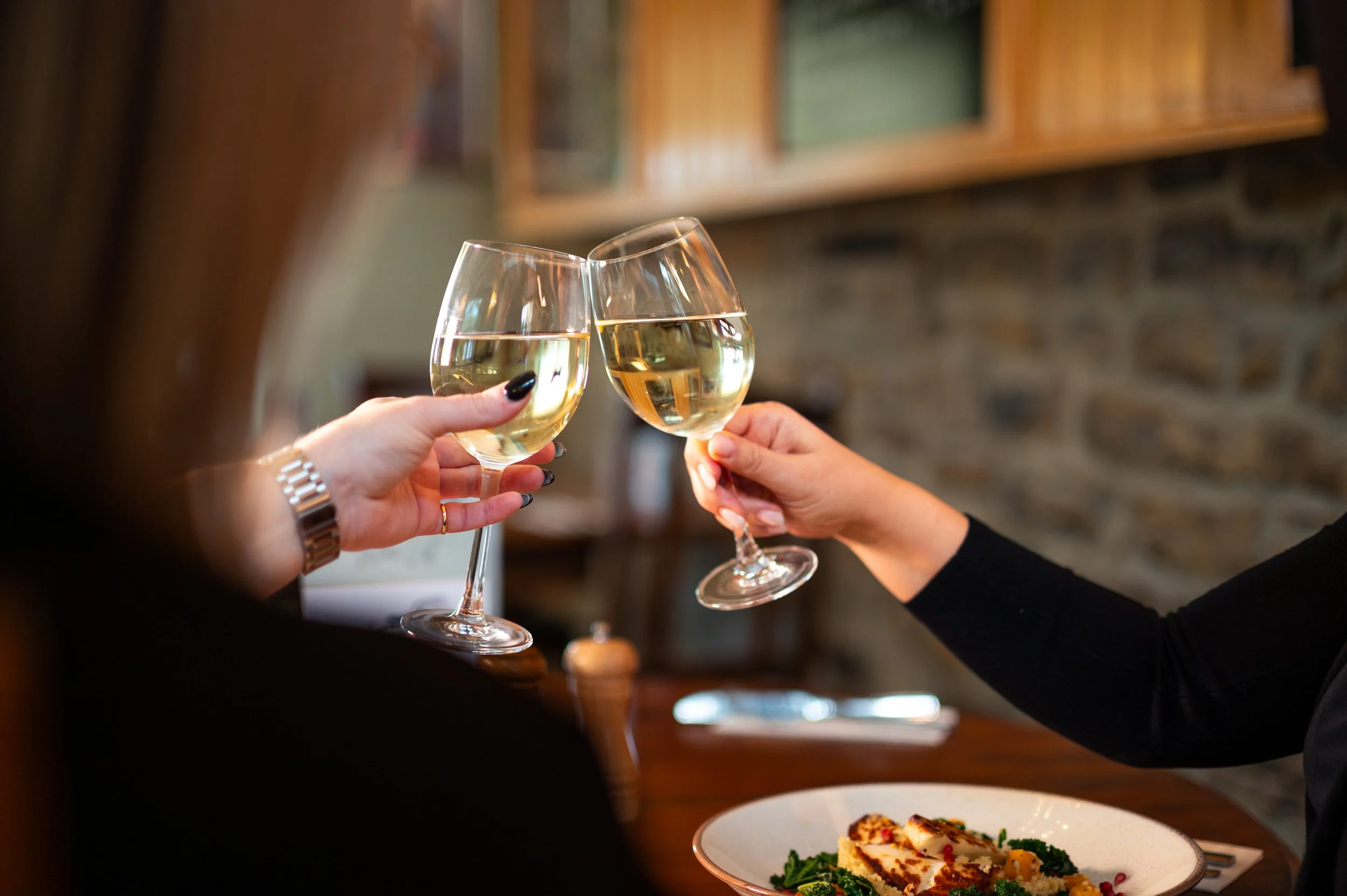 Two women clinking glasses of white wine at a dinner table in a cozy restaurant setting.