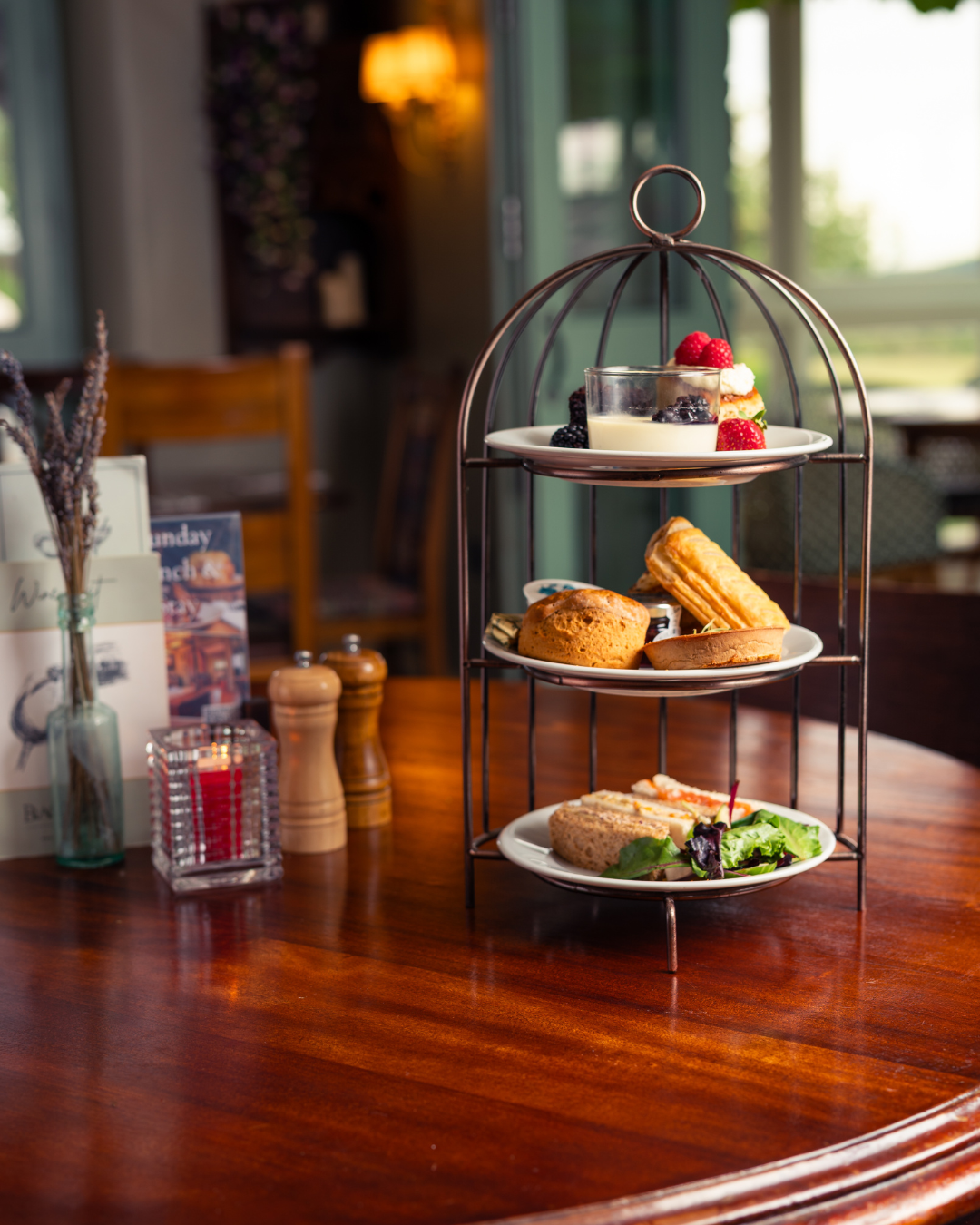 hree-tiered dessert stand with assorted desserts including cake slices, cookies, jam, and finger sandwiches on a wooden table