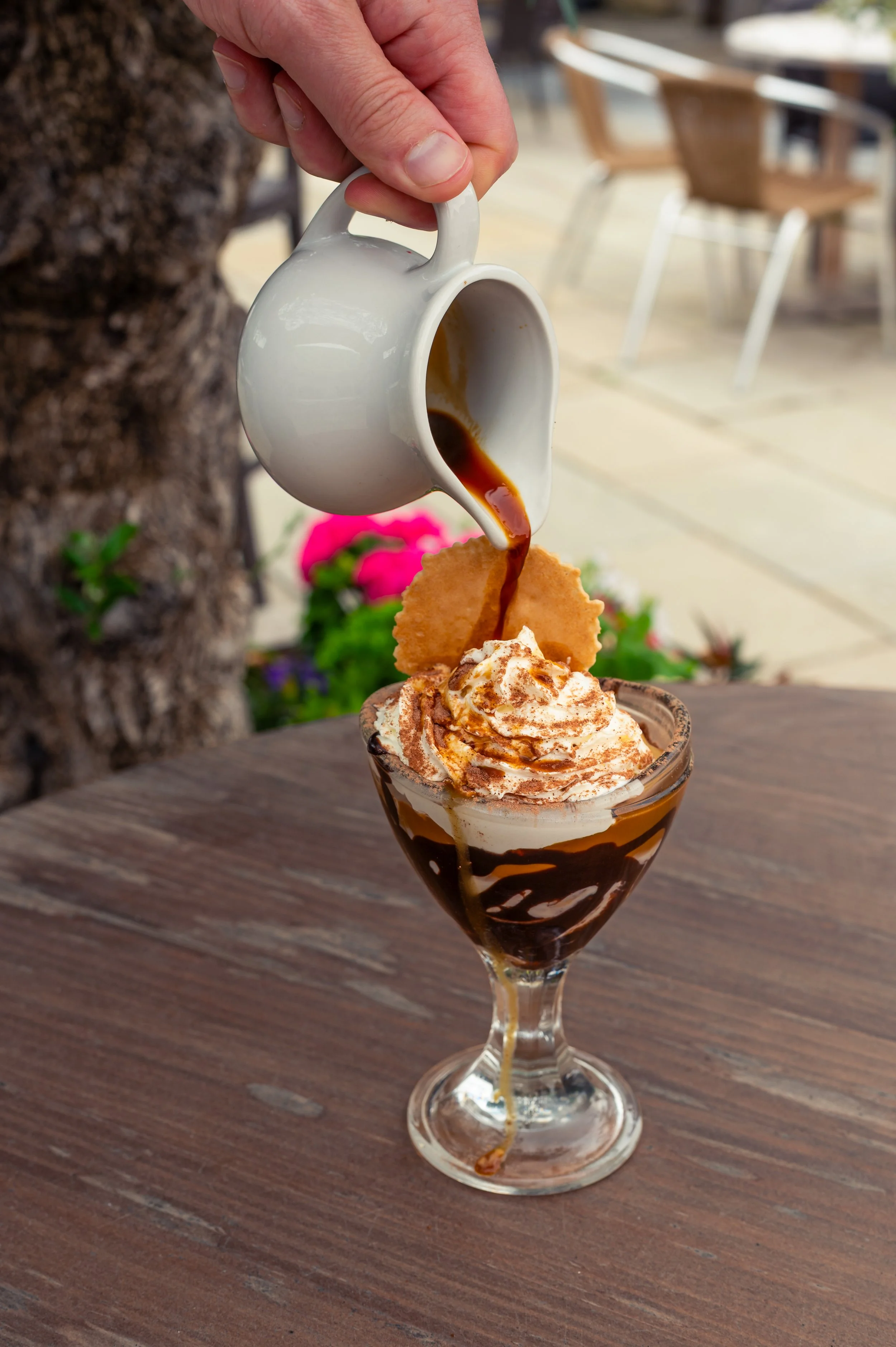 Hand pouring coffee over whipped cream and caramel in a glass dessert cup with a cookie on top, set on a wooden table outdoors.