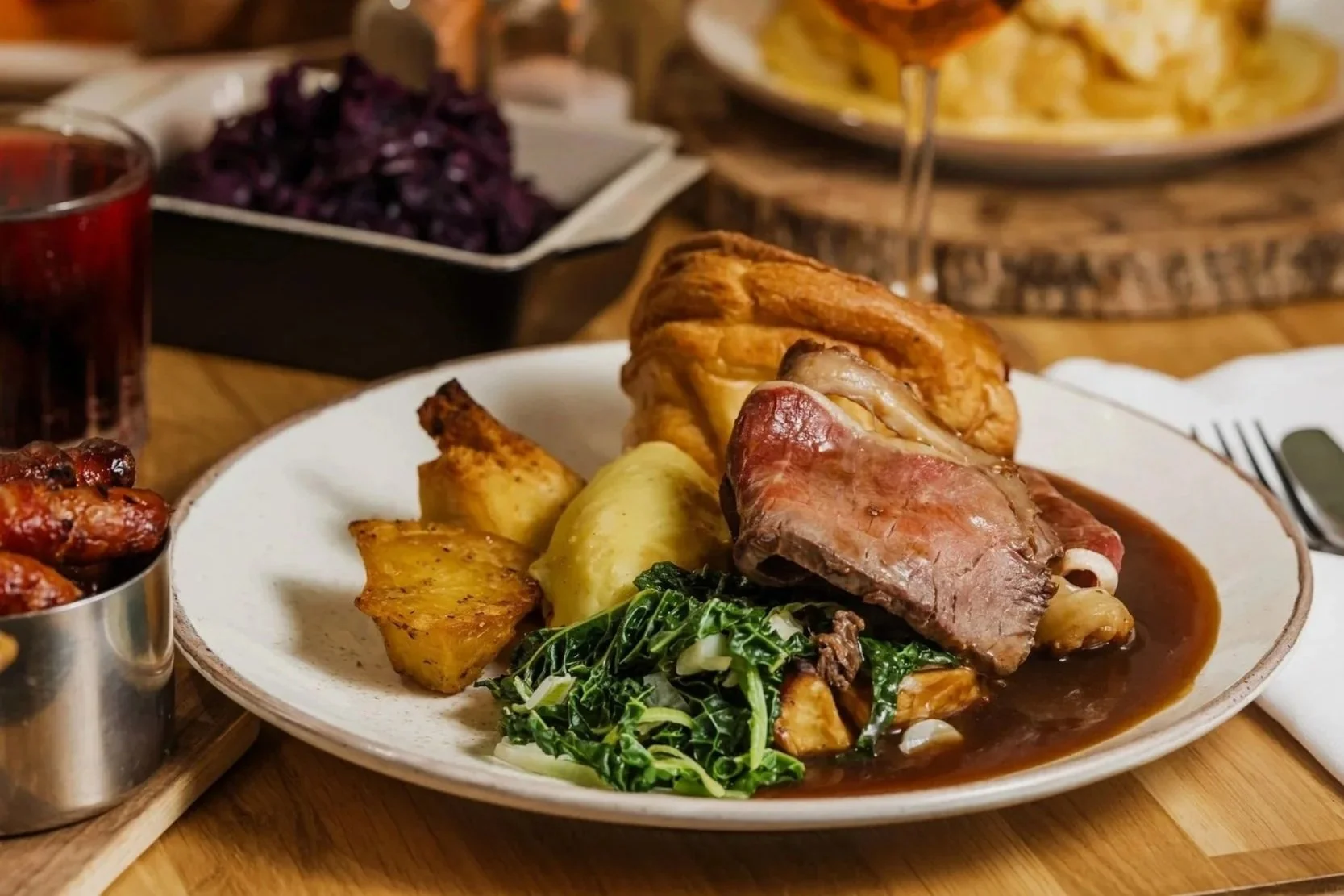 A plate of American-style roast beef with gravy, mashed potatoes, roasted potatoes, sautéed greens, and a piece of fried chicken, with sides of coleslaw, a glass of red wine, and other dishes in the background.