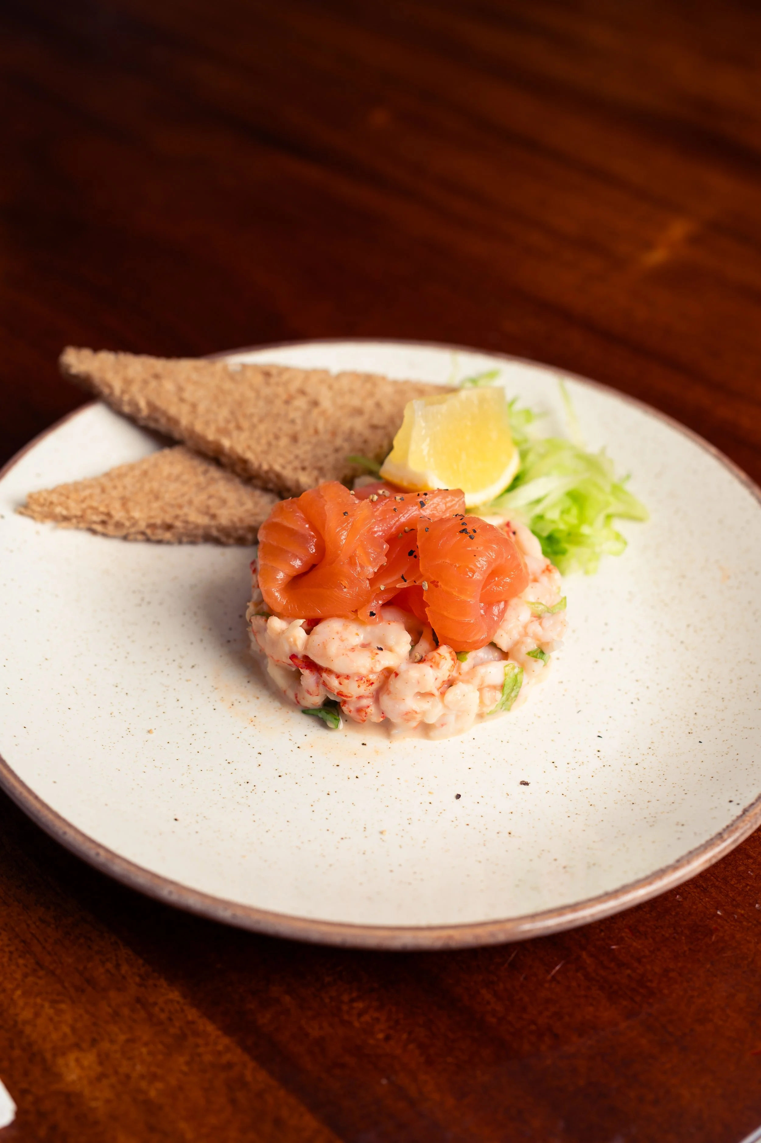 A plate with smoked salmon on lettuce, lemon wedge, and two slices of toast on a wooden table.