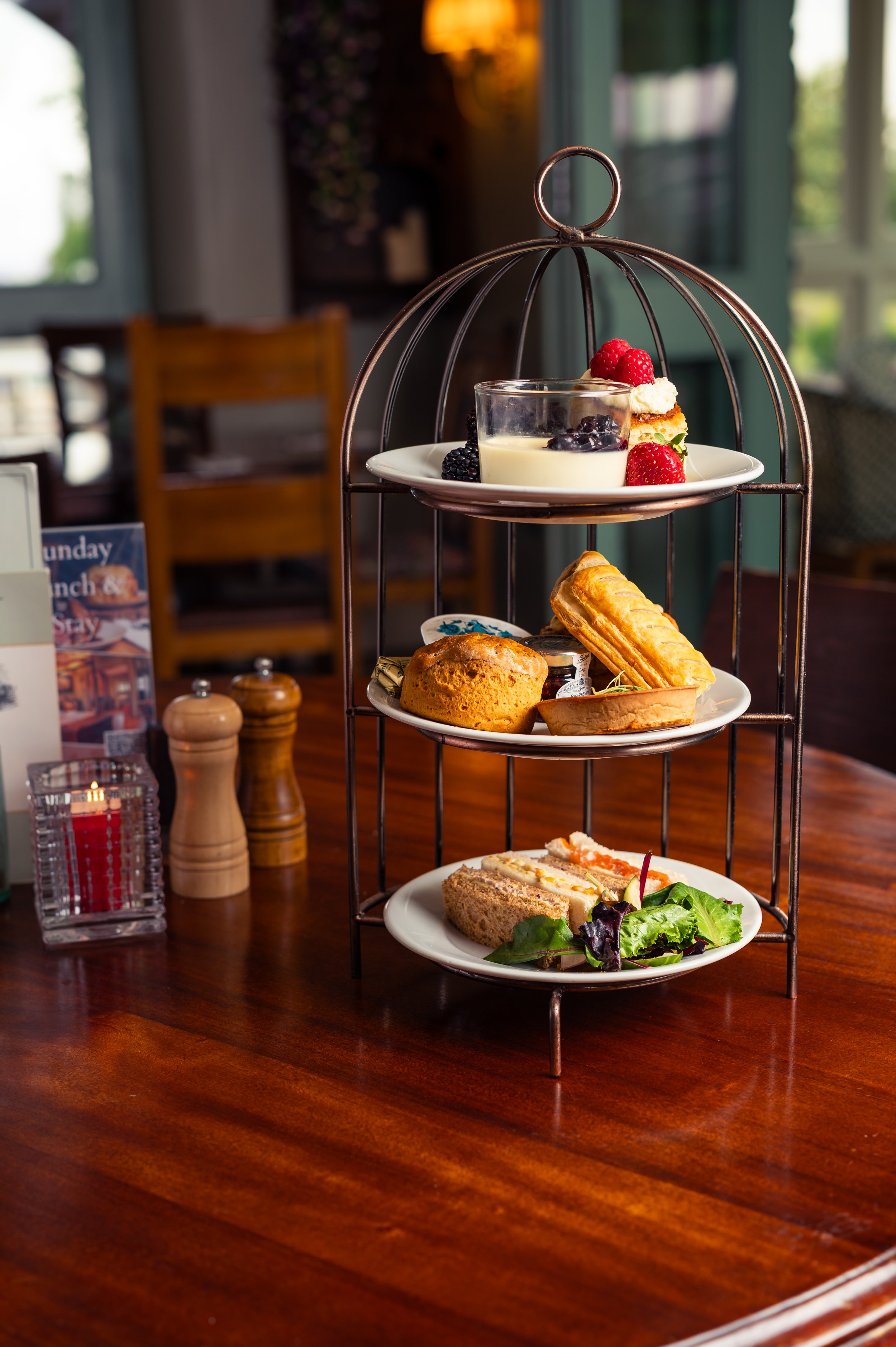 Three-tiered serving tray with assorted breakfast items, including a glass of yogurt with fruit, pastries, a sandwich, and a salad, on a wooden table in a cozy restaurant setting.
