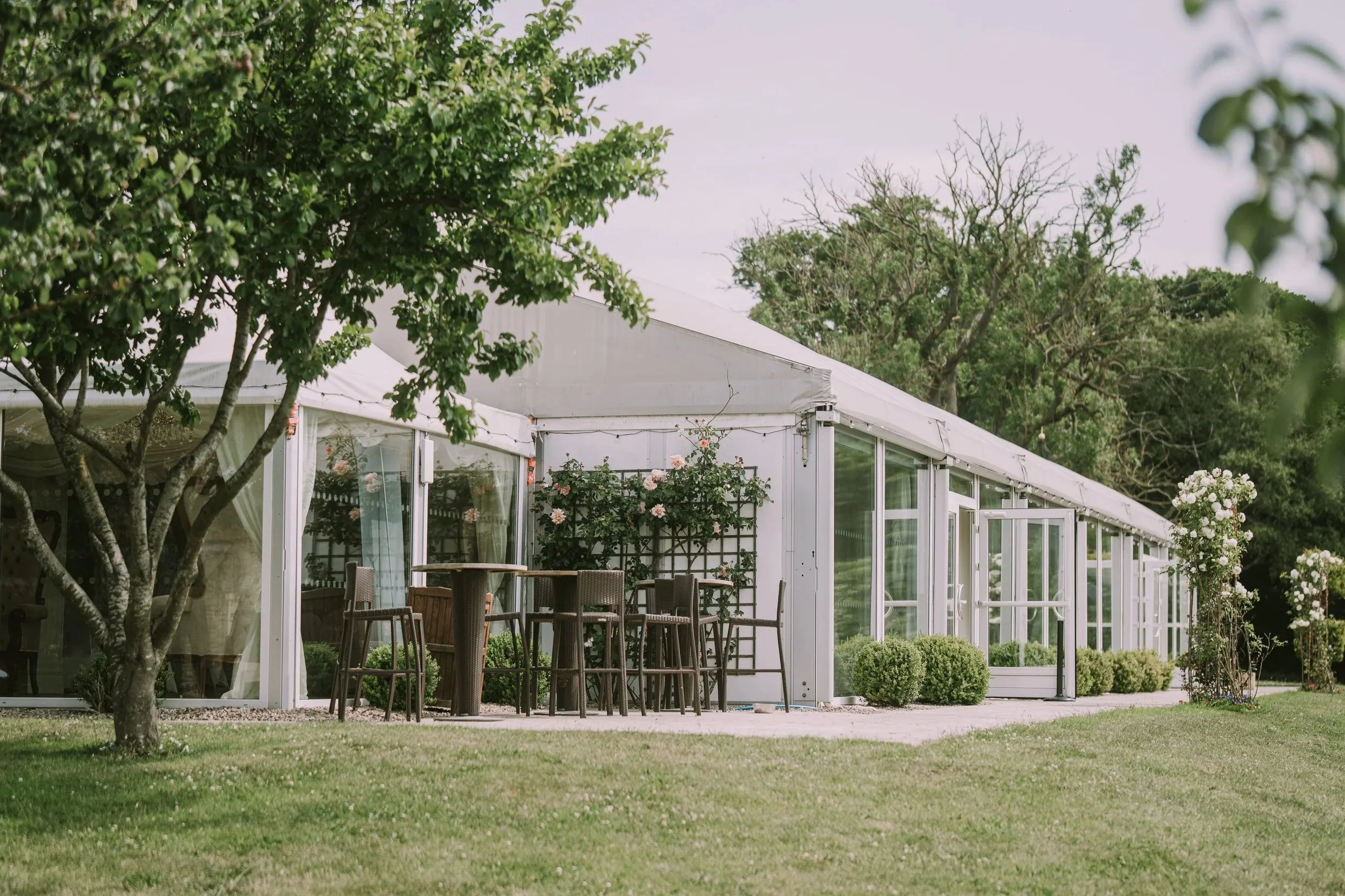 A glass greenhouse surrounded by greenery and blooming white and pink flowers, with a lawn and outdoor seating area.