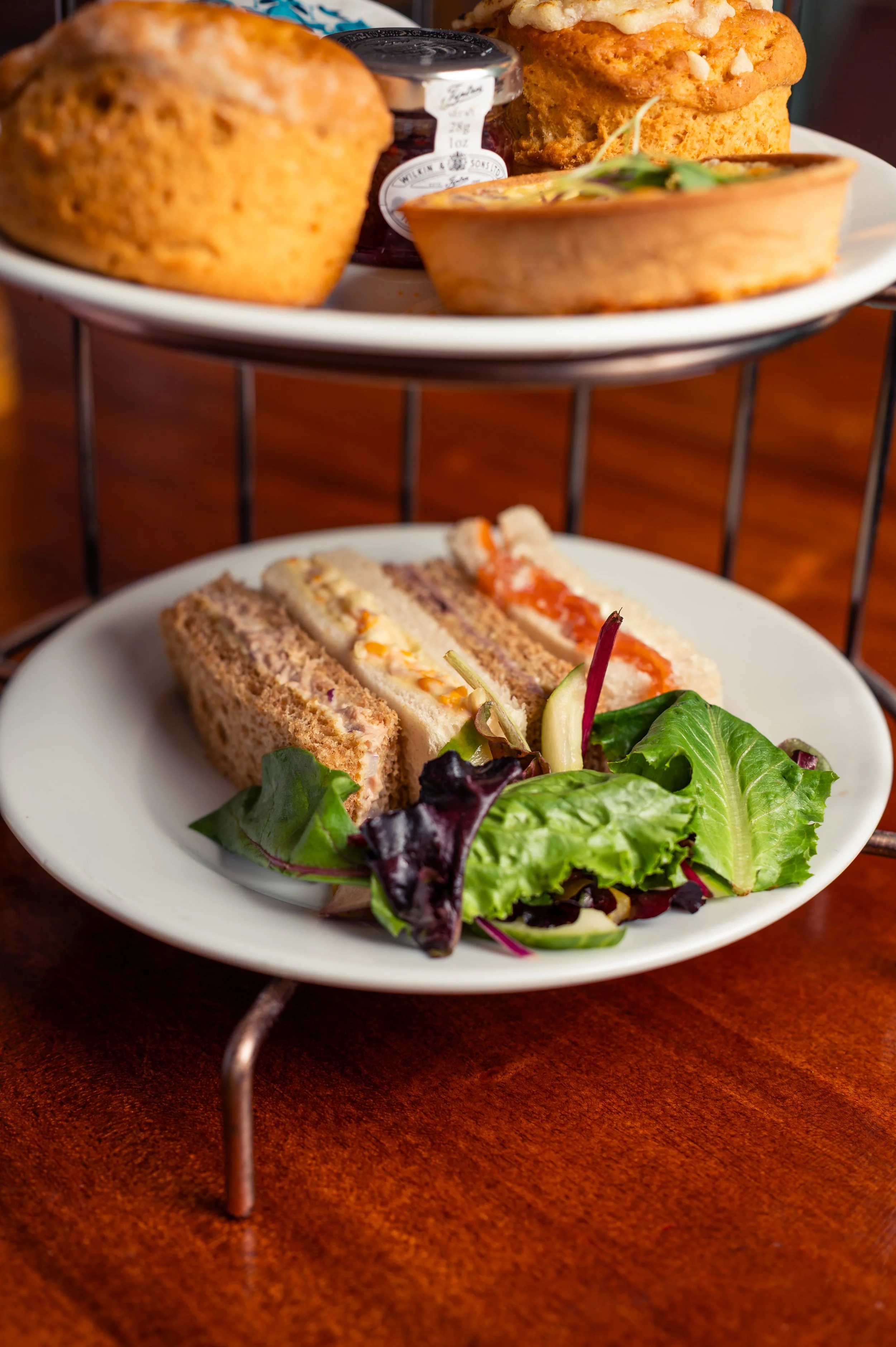 A two-tiered serving tray with an assortment of foods. The top tier includes a loaf of bread, a small jar of jam, a slice of savory pie, and a piece of rich cake. The bottom tier features a small salad with mixed greens, slices of finger sandwich wit