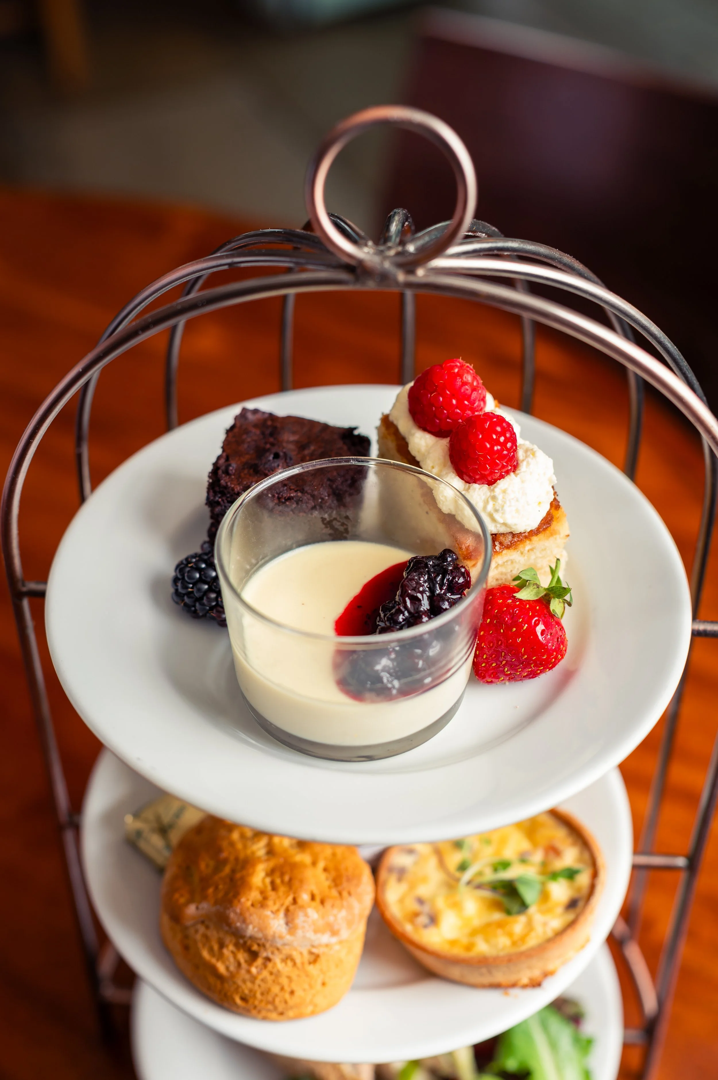 A three-tiered serving tray with assorted desserts: a piece of chocolate cake with blackberries, a small cake topped with whipped cream and raspberries, a shot glass of custard with berry compote, and a strawberry. The tray has a metal handle, and there is a plate of food below.