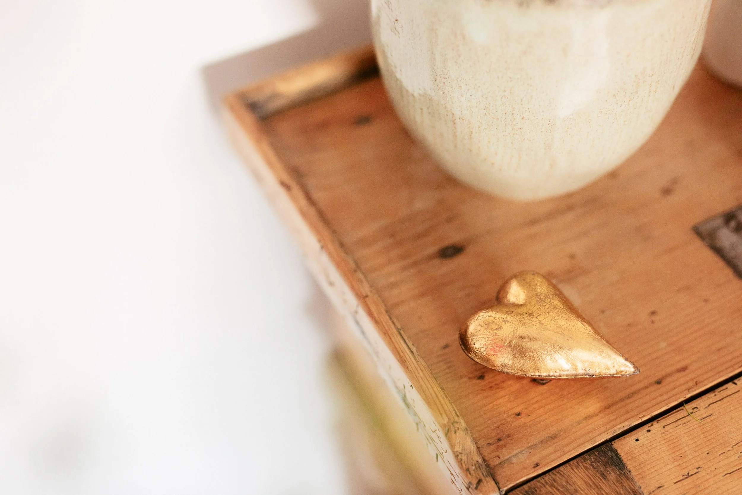 Ein goldfarbenes Herz liegt auf einem Holztisch neben einer weißen Vase.