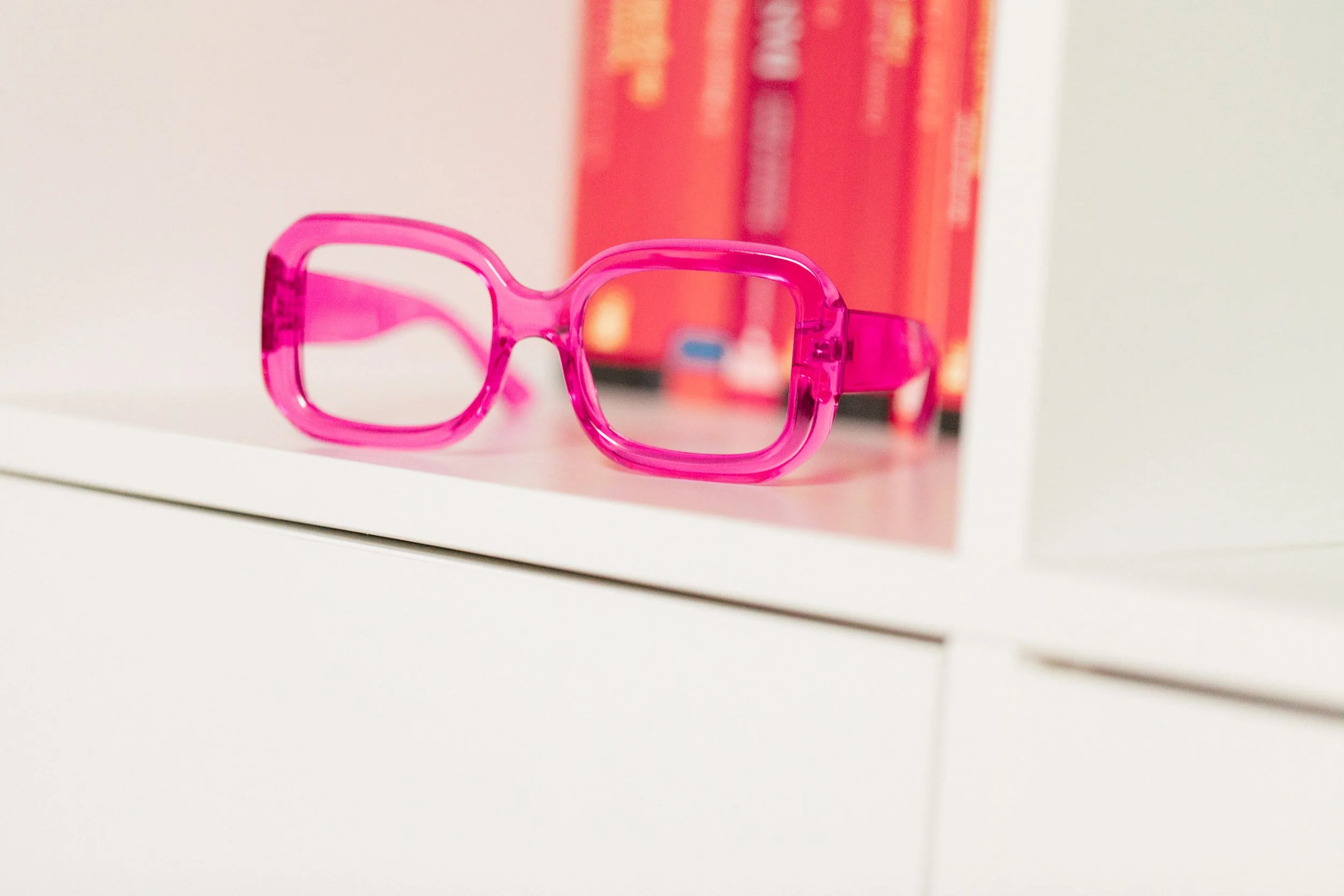 Bunte pinke Brille auf weißem Regal, mit roten Büchern im Hintergrund.