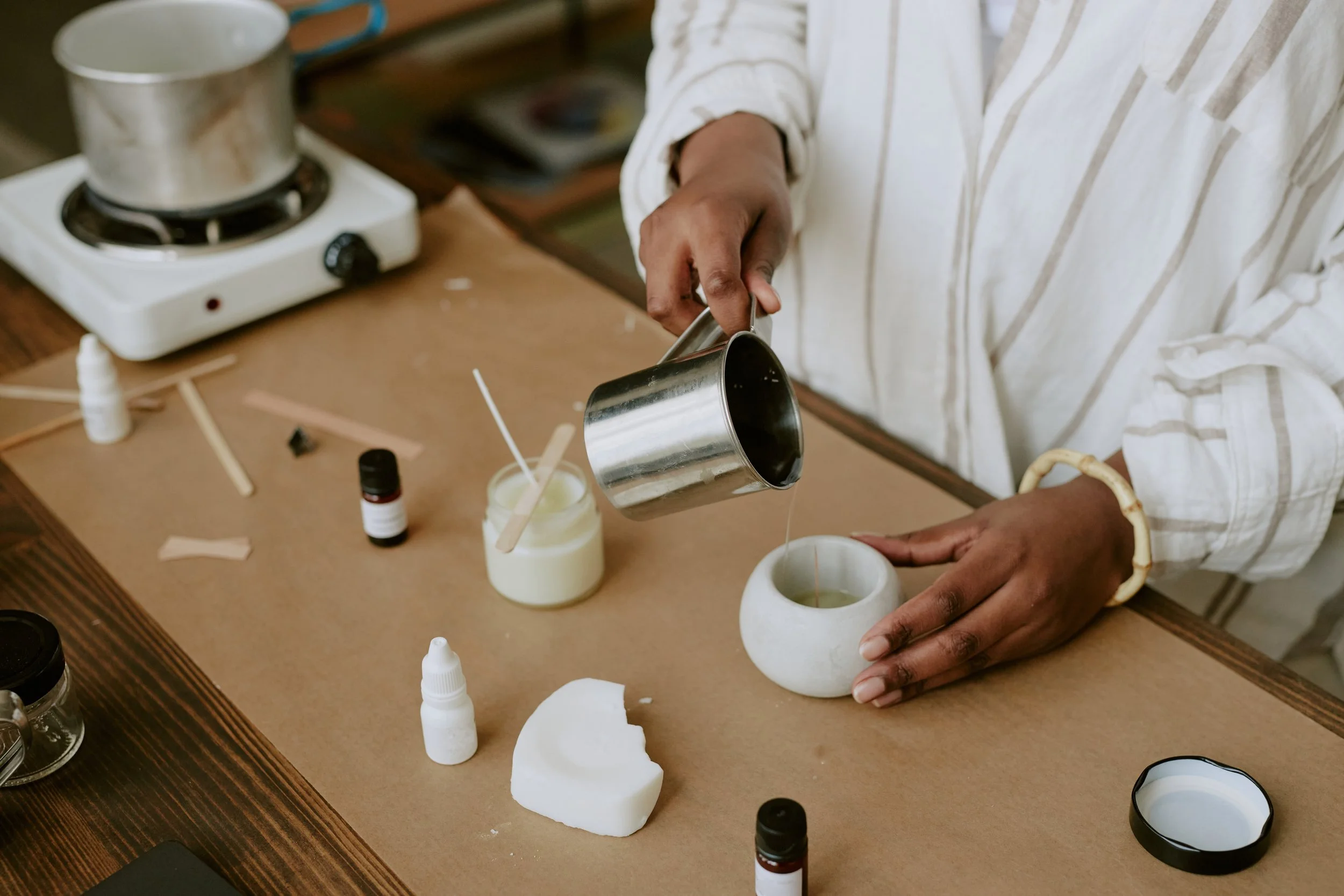 Une personne verse de la cire chaude dans une petite coupe en pierre sur une table de bois lors d'un atelier de création de bougies, entourée de divers outils et contenants pour la fabrication de bougies.