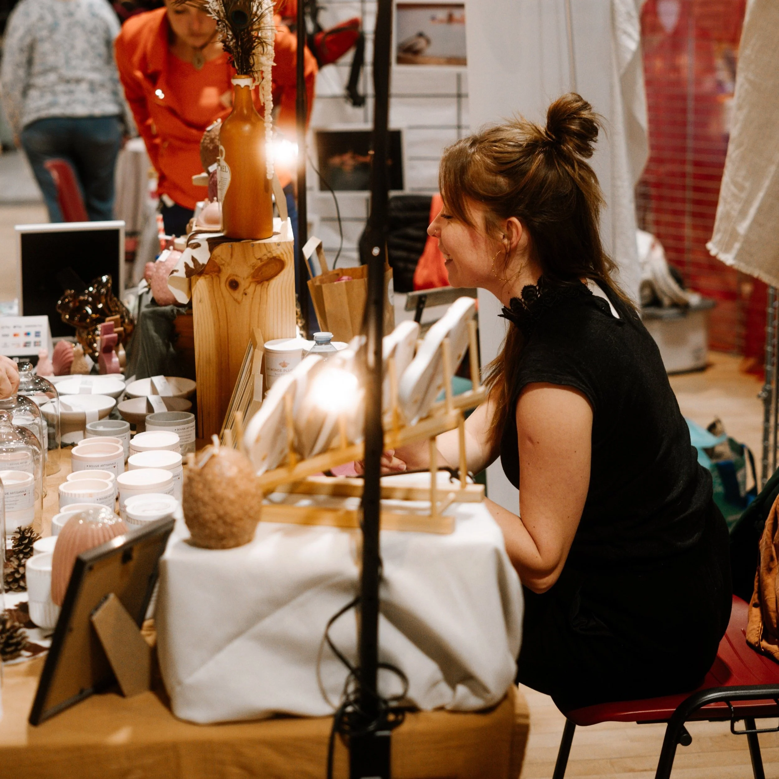 Femme souriante à un stand de marché ou marché artisanal, entourée de produits décoratifs et artisanaux.