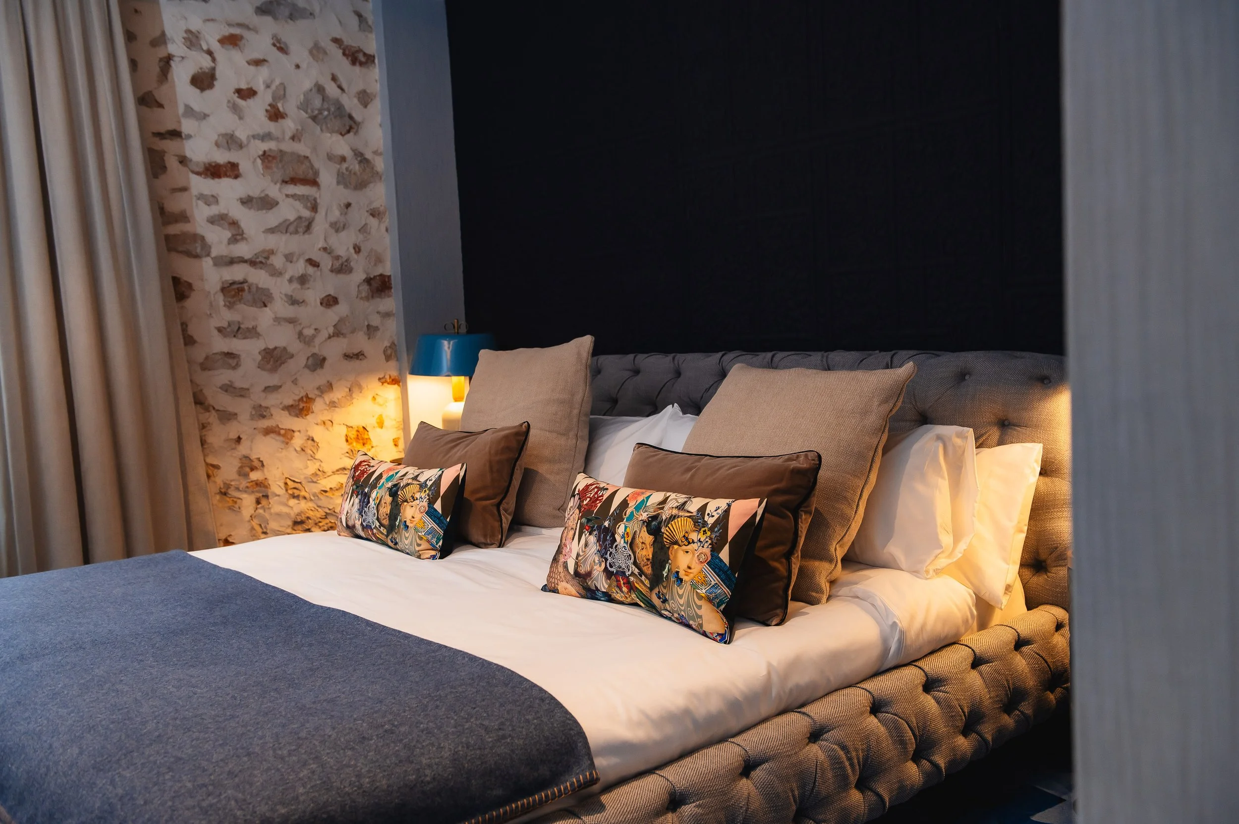 Cama con varios cojines y decoración moderna en una habitación con pared de piedra y cortinas beige.