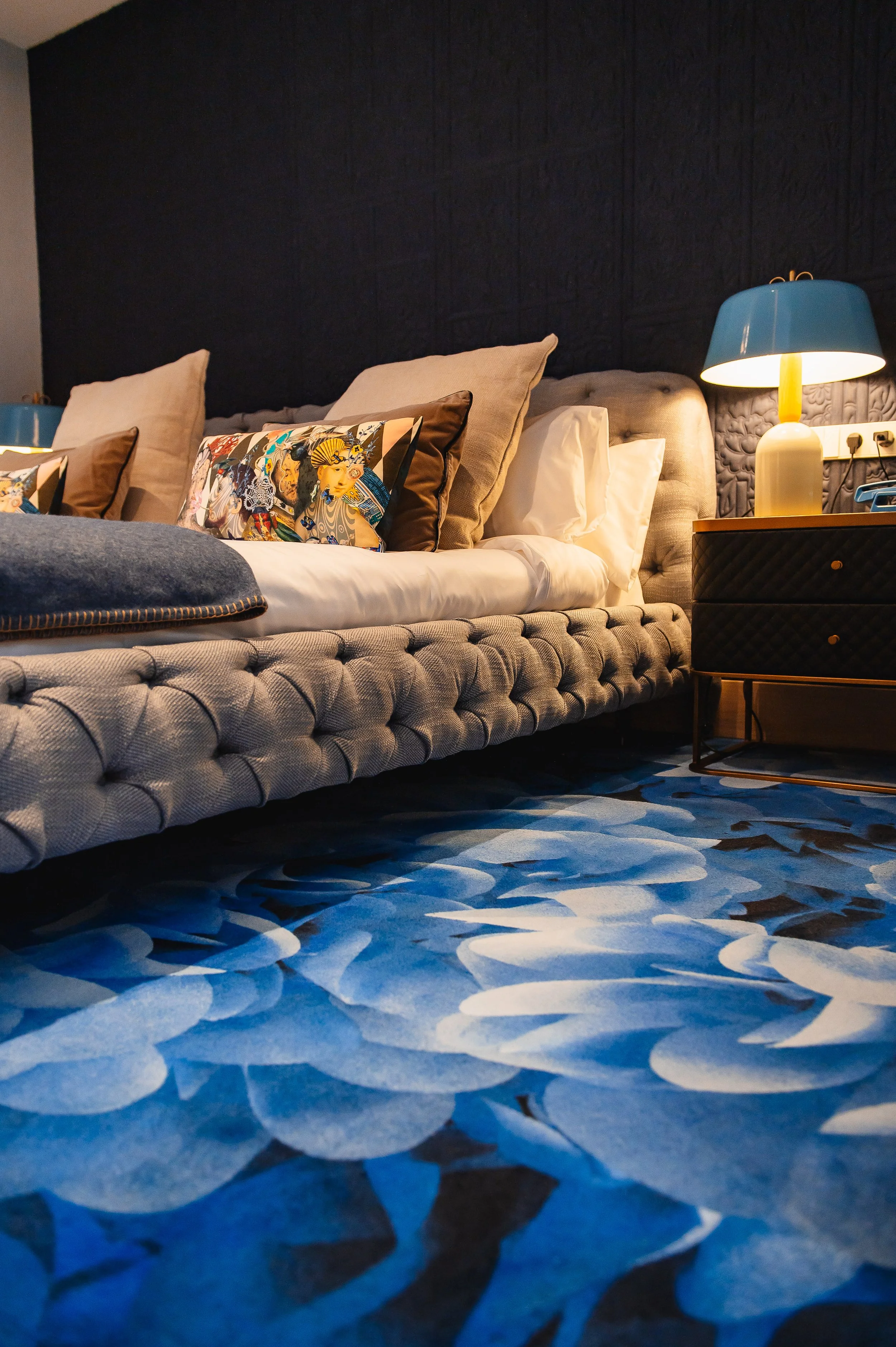 Habitación moderna con cama con cabecera acolchada gris, varias almohadas, una lámpara de mesa azul y blanca, y una alfombra azul con patrón de hojas blancas y negras.