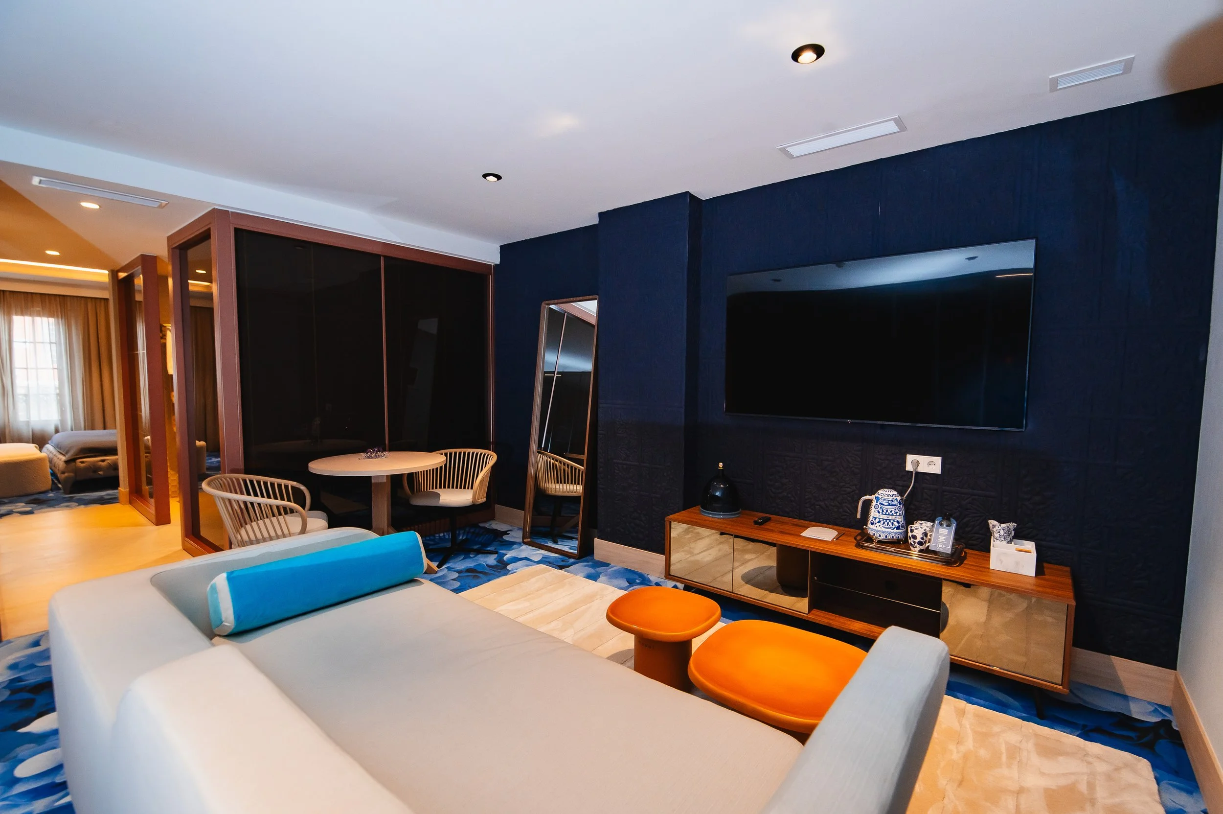 Sala de estar moderna con sofá blanco, dos ottomans naranjas, un mueble de madera con objetos decorativos y un televisor grande en la pared de fondo.