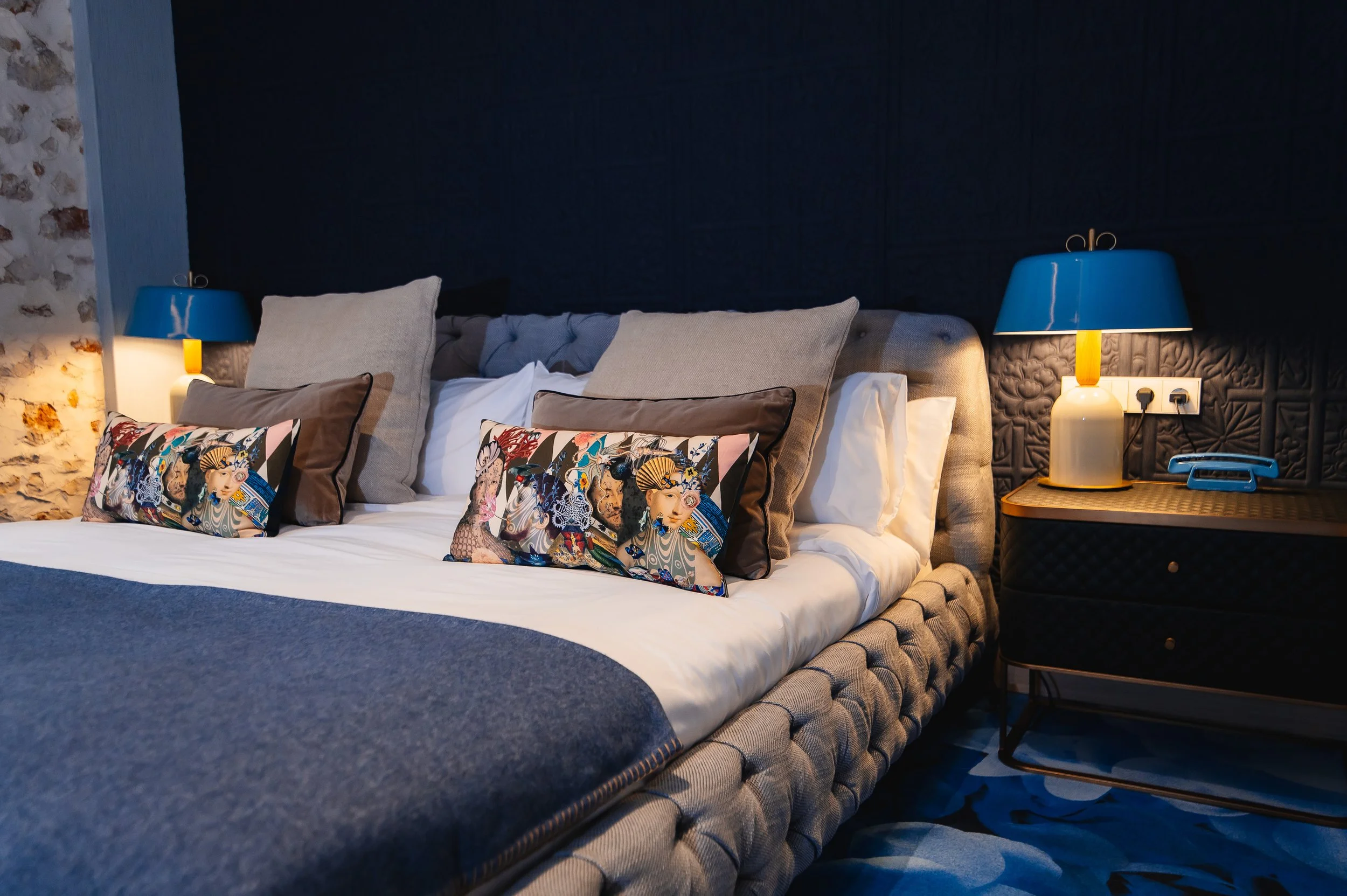 Cama de matrimonio con varias almohadas y cojines, lámparas azules y una mesita de noche con teléfono y lámpara en una habitación decorada con paredes de piedra y estilo moderno.