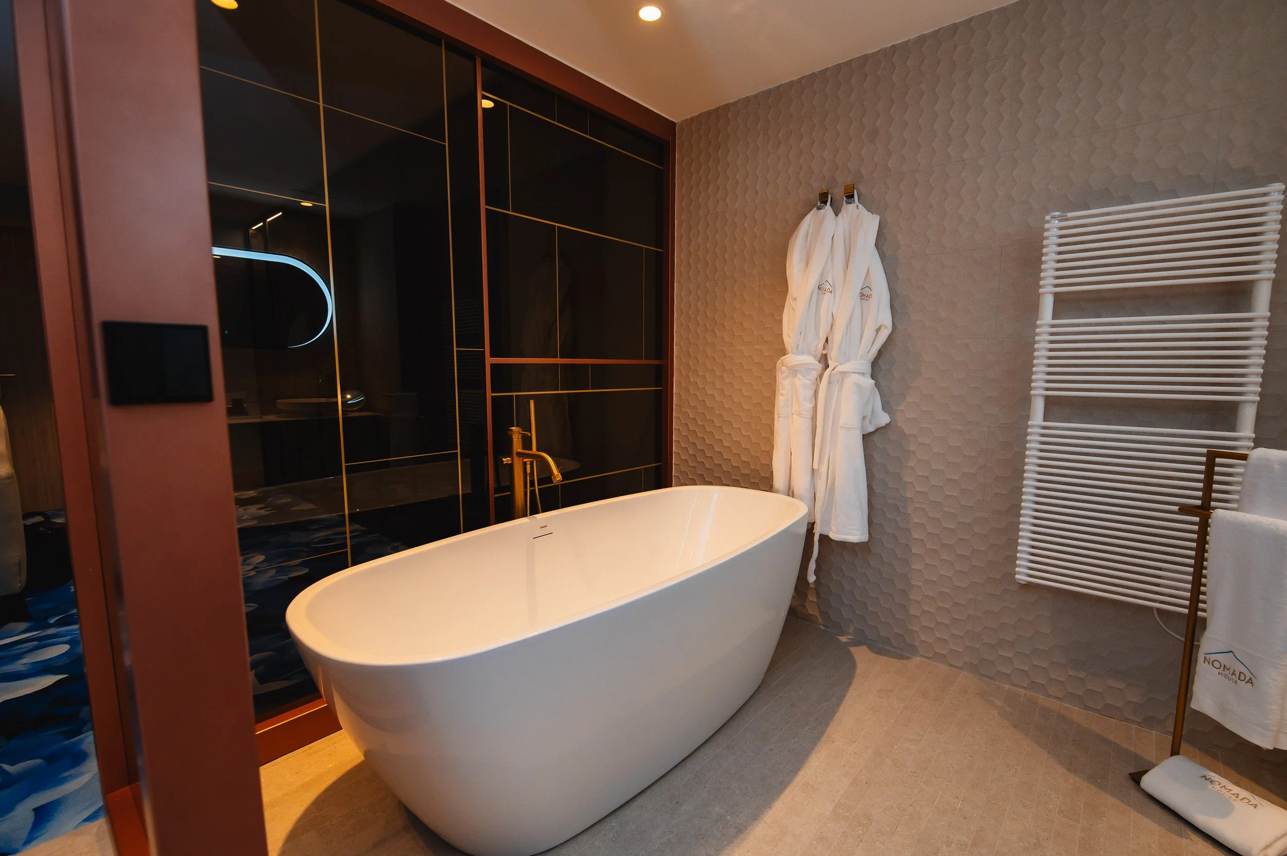 Baño moderno con bañera de pie, toallas blancas en colgador y radiador, pared decorativa y mampara de vidrio.