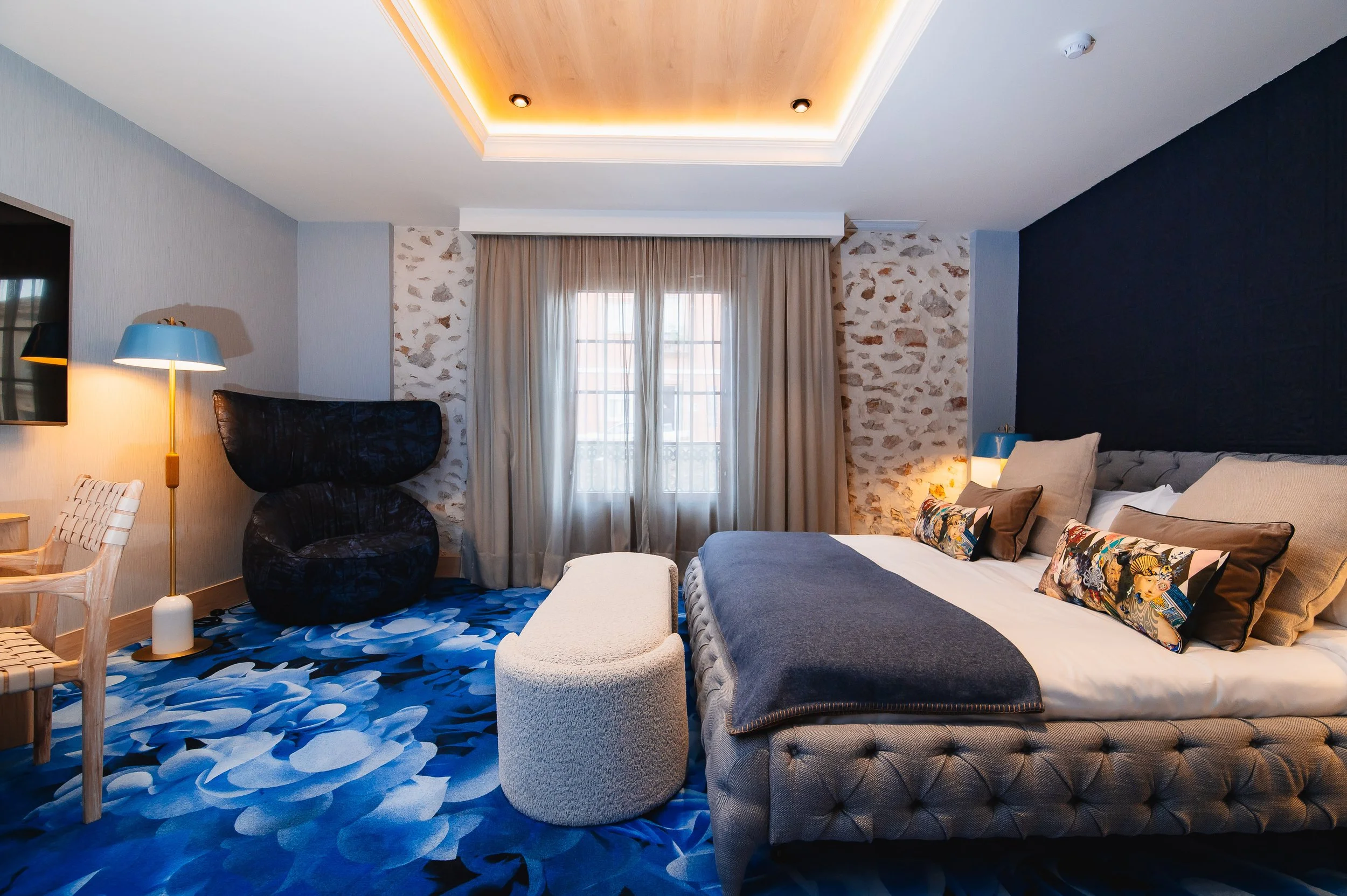 Habitación de hotel con cama grande, sillas, sillón azul y cortinas. Piso con alfombra azul con patrones, paredes de piedra y pintura blanca. Iluminación cálida.