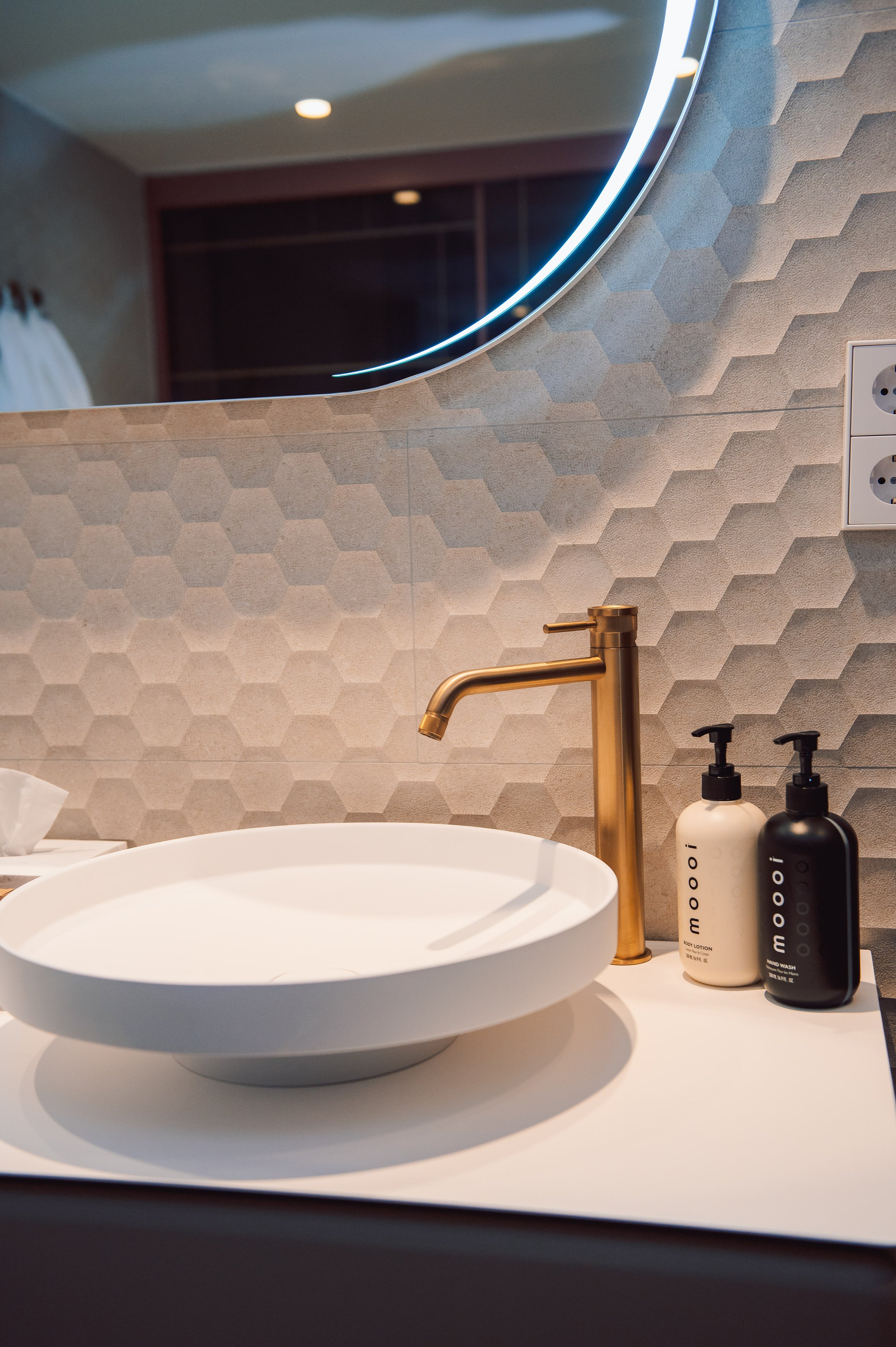 Lavabo con jabón y loción en un baño moderno con pared de azulejos hexagonales y un espejo redondo con iluminación LED.
