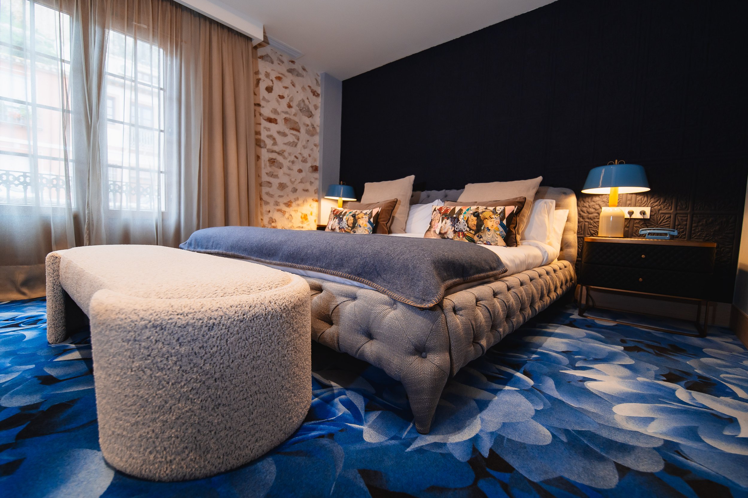 Habitación de hotel con cama grande, almohadas decorativas, lámparas de mesa azules, cortinas beige y una alfombra azul con patrón floral.