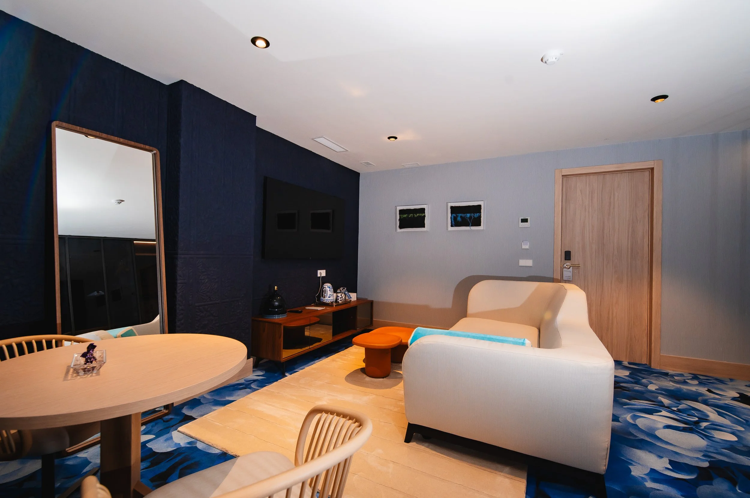 Sala de hotel con sillón beige, mesa ovalada de madera, espejo grande, televisión en la pared negra, pintura en la pared gris, puerta de madera, alfombra con patrón azul, muebles de madera y paredes azules y grises.
