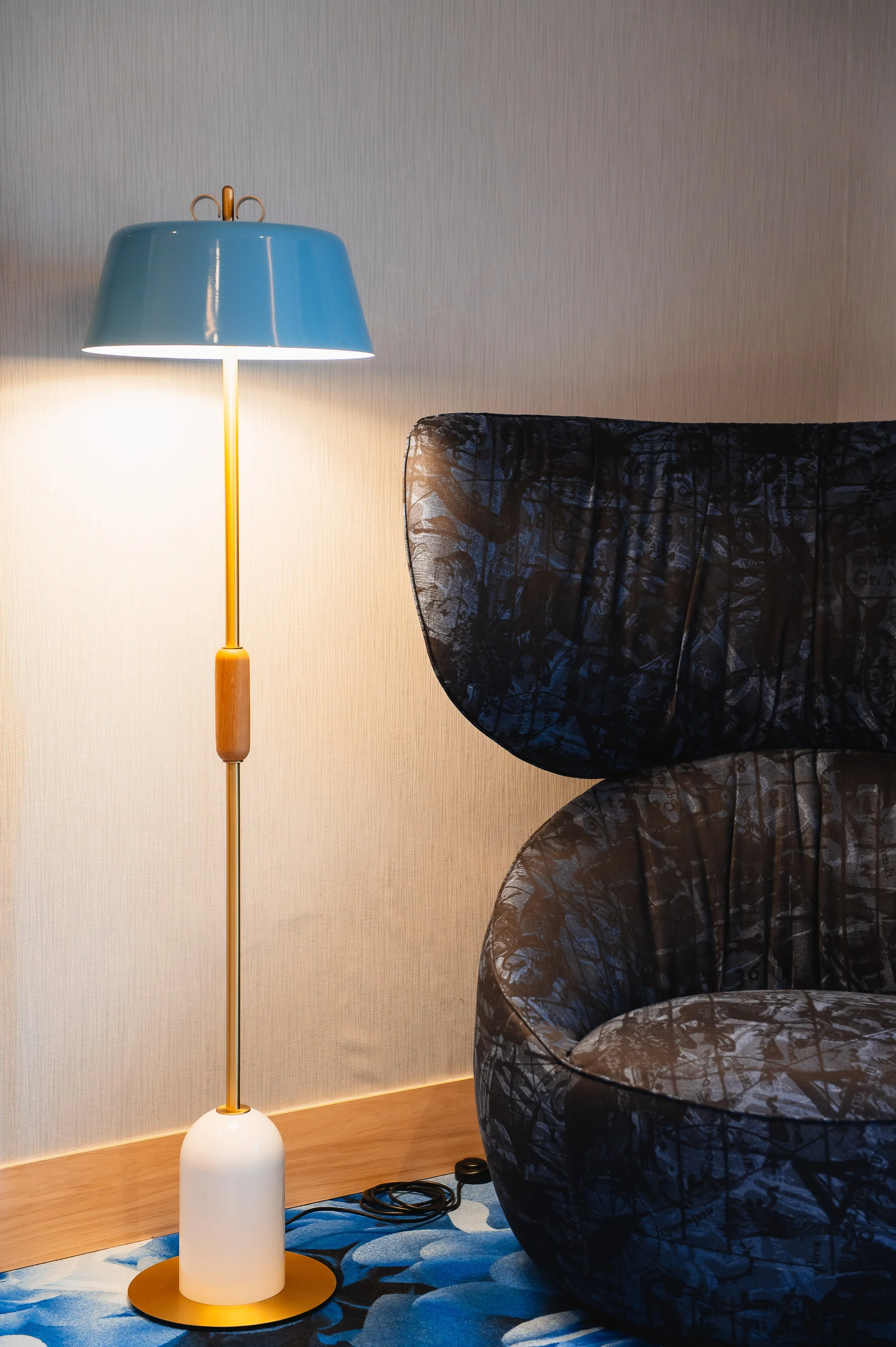 Lámpara de pie junto a una silla cubierta con tela negra y patrones en una habitación con paredes beige y alfombra con patrones en tonos azules.