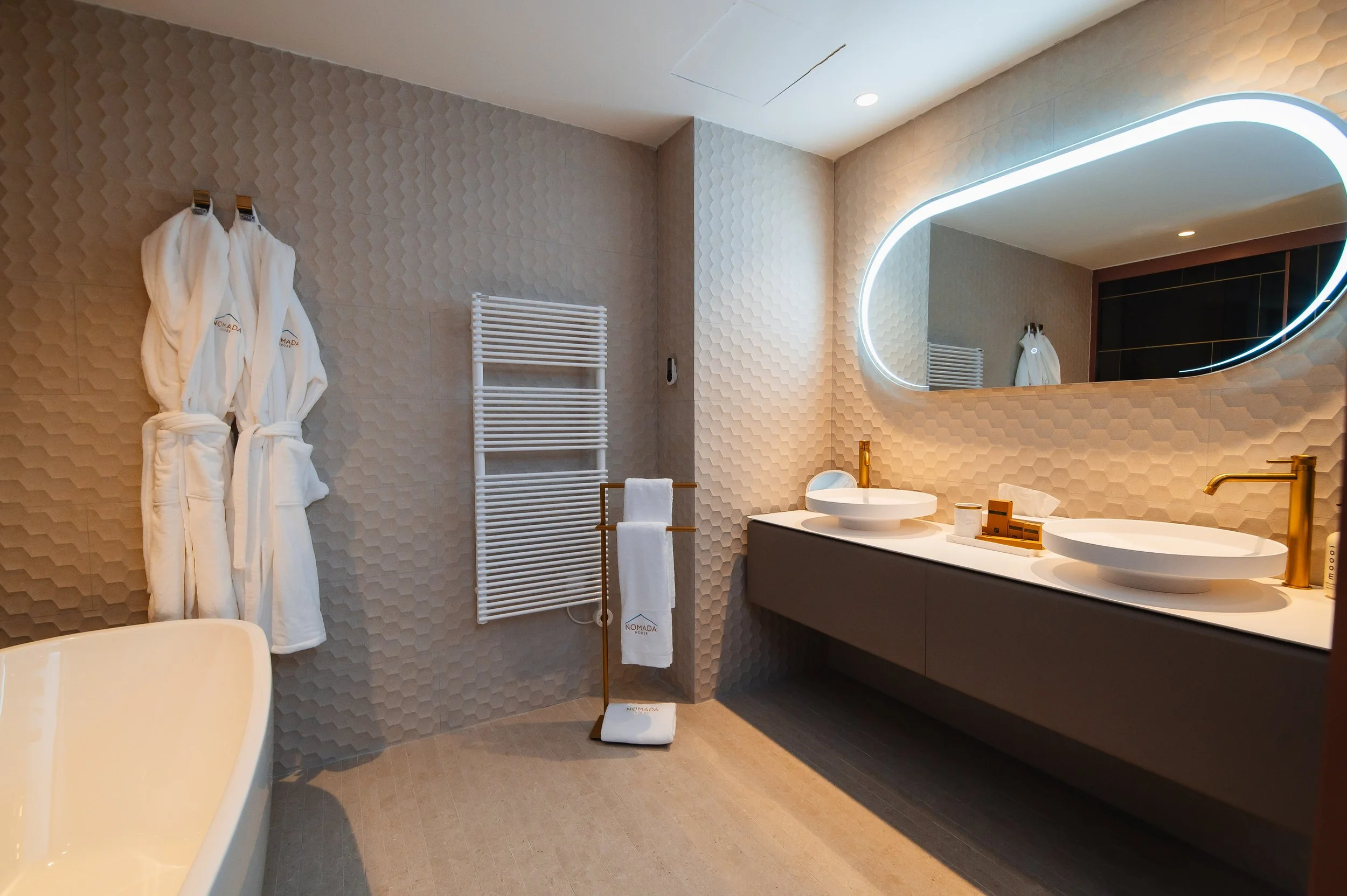 Baño moderno con dos lavabos redondos en un mueble gris oscuro, gran espejo ovalado con iluminación LED, toallas blancas en una estructura de madera y una bata blanca colgada en la pared.