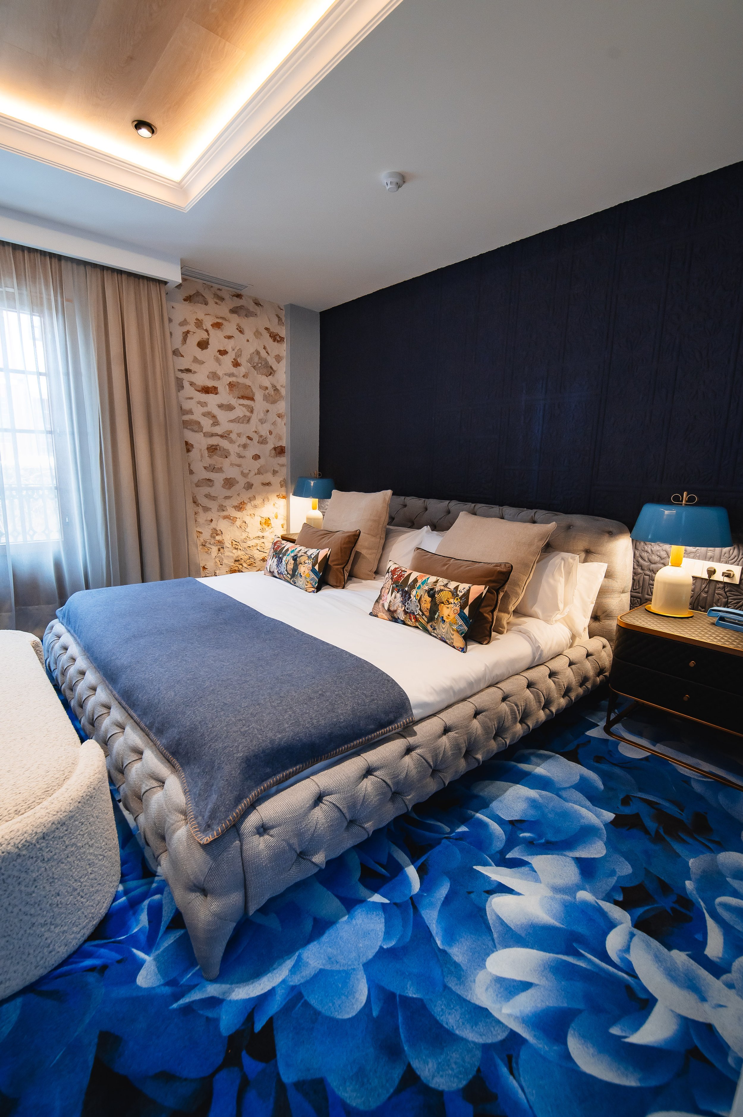 Habitación decorada con una cama grande con cabecera acolchada, varias almohadas decorativas, una manta gris y sábanas blancas. A la izquierda hay cortinas beige y al fondo, una pared de piedra y una pared azul. A la derecha hay mesas de noche con lá
