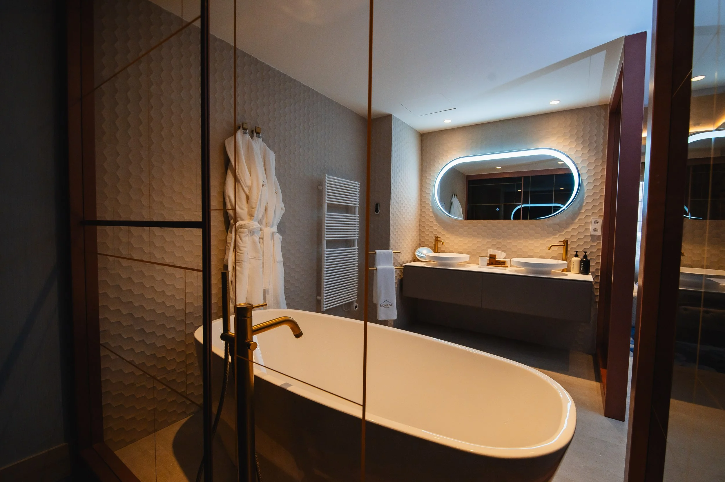 Baño moderno con bañera de pie, espejo con iluminación de neón, lavabos dobles con dorados, paredes decoradas con azulejos en relieve, toallas blancas, ducha y accesorios en colores oscuros.