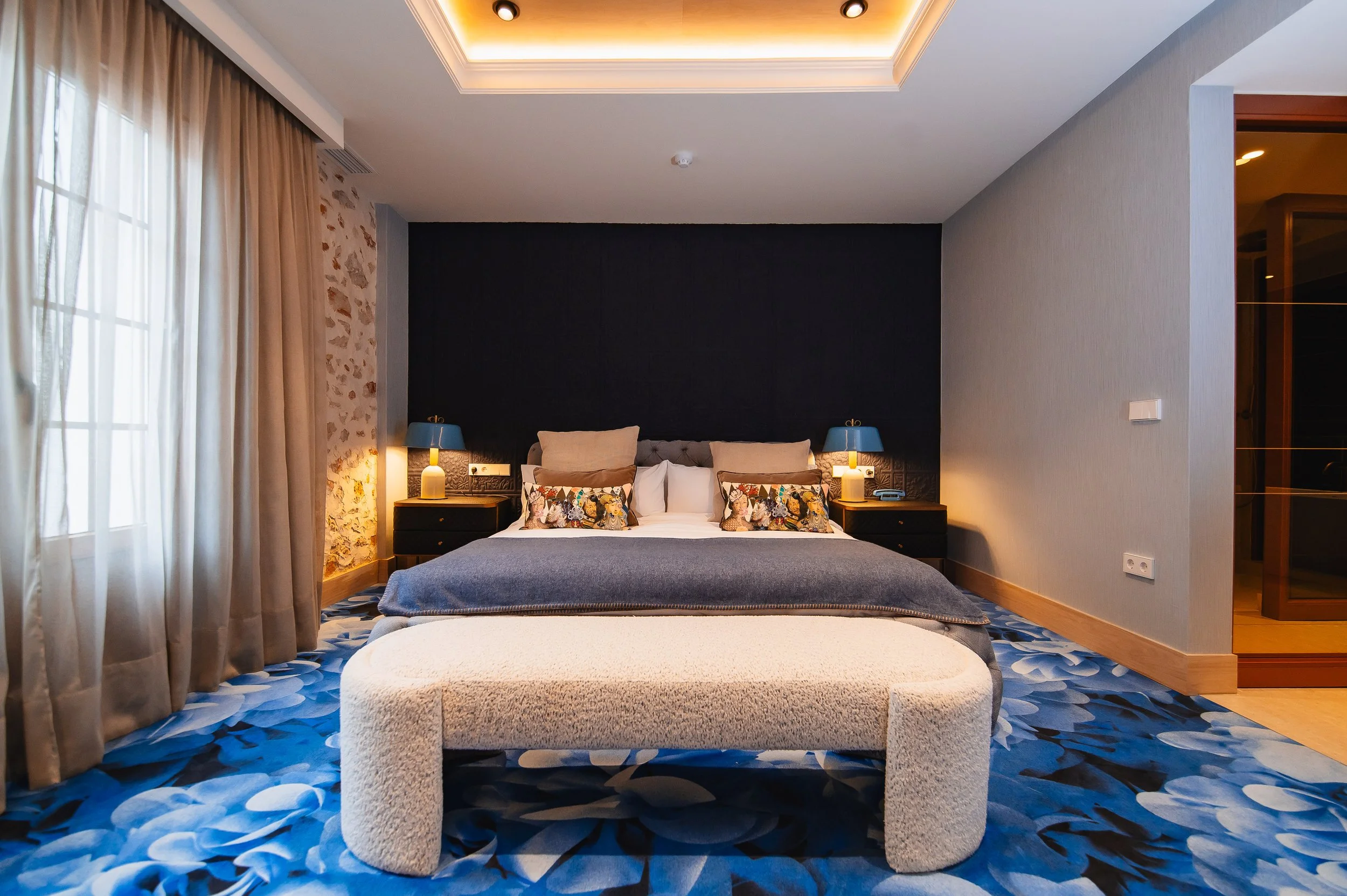 Habitación de hotel con cama grande, dos mesitas con lámparas azules, cortinas beige y alfombra de diseño azul