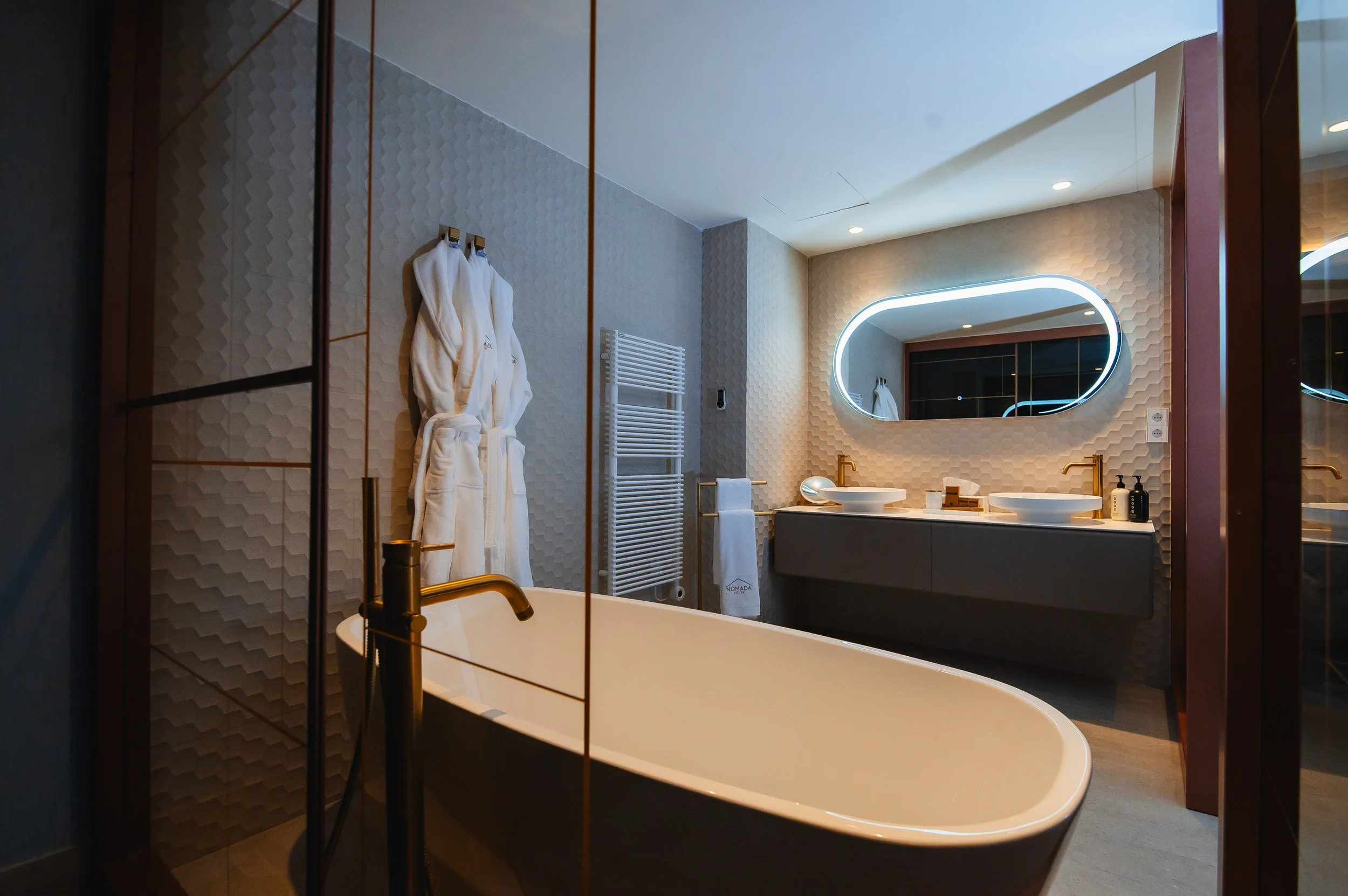 Baño moderno con bañera de pie, espejo de luz, lavabos dobles con grifos dorados, y rutina de toallas blancas, en un diseño elegante y minimalista.