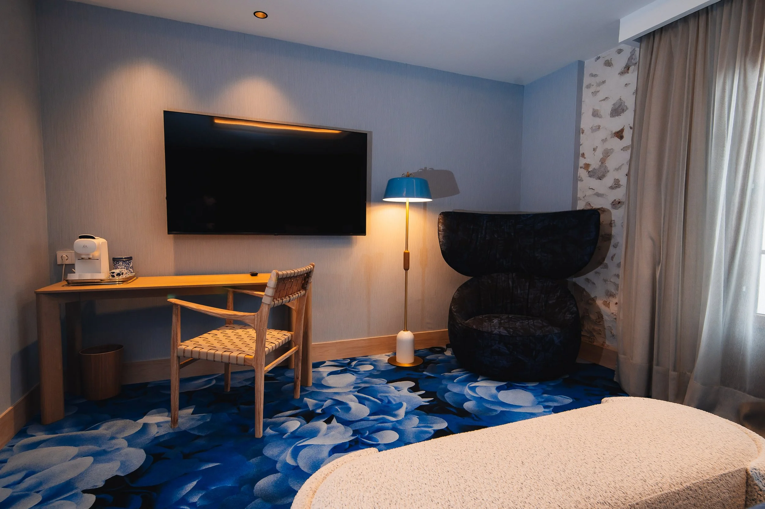 Habitación de hotel con televisor, lámpara de pie, silla, escritorio y cortinas.