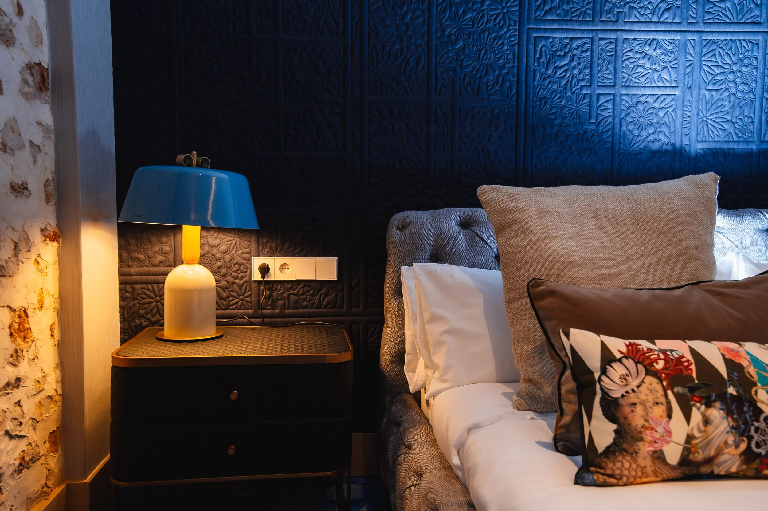 Dormitorio con cama, cojines decorativos, lámpara azul sobre una mesa negra, pared negra con textura decorativa, enchufe eléctrico a la pared, pared de ladrillos a la izquierda.