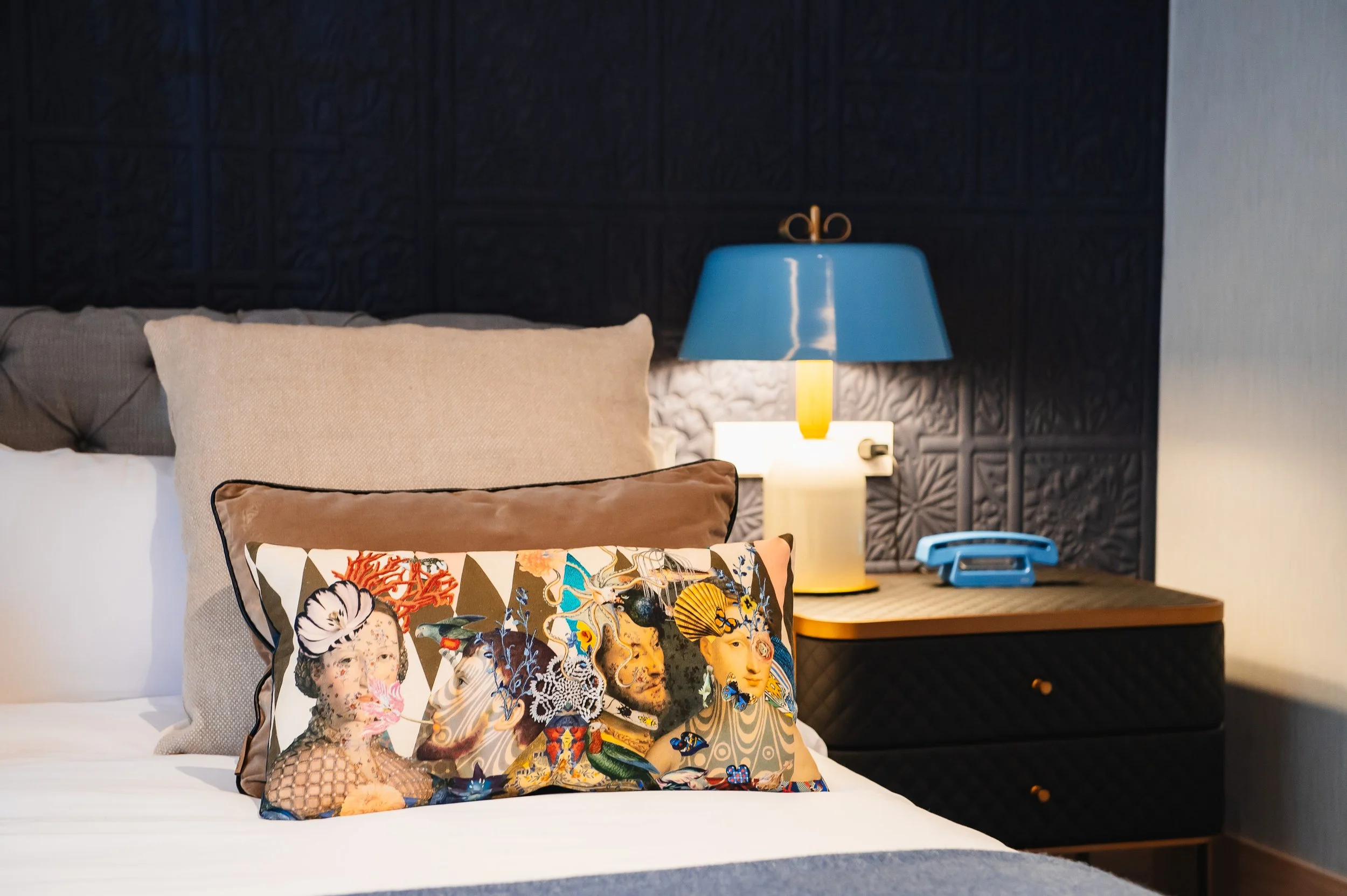 Cama con almohadas decorativas y lámpara azul en la mesita de noche, en una habitación moderna con paredes oscuras.
