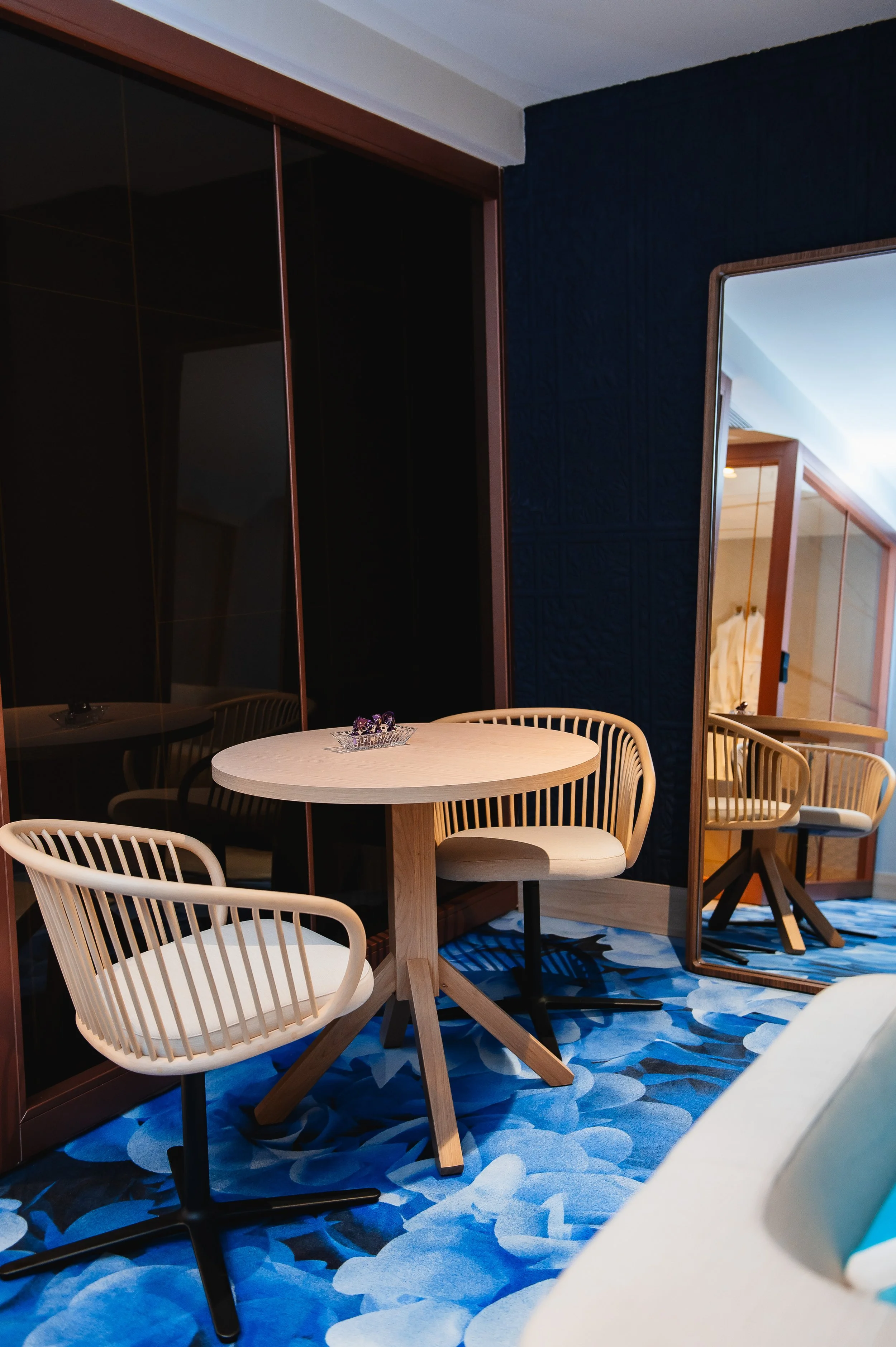 Un mueble con sillones de mimbre y una mesa de madera en una habitación con alfombra azul con patrones blancos y un espejo de cuerpo entero.