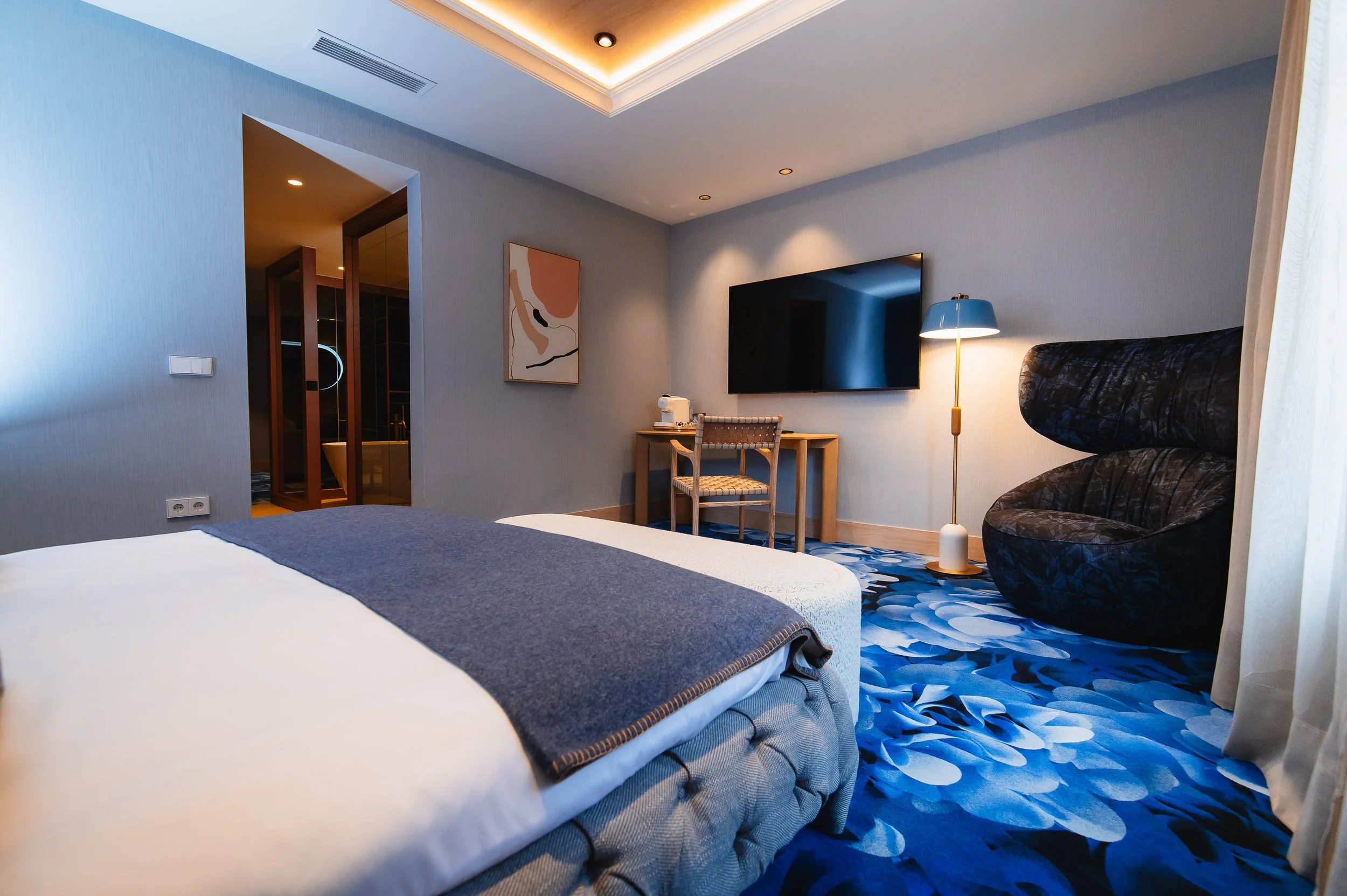 Habitación de hotel con cama grande, pared azul, alfombra azul con patrón abstracto, silla de mimbre, mesa de escritorio con televisión y lámpara, y un área de baño visible en el fondo.