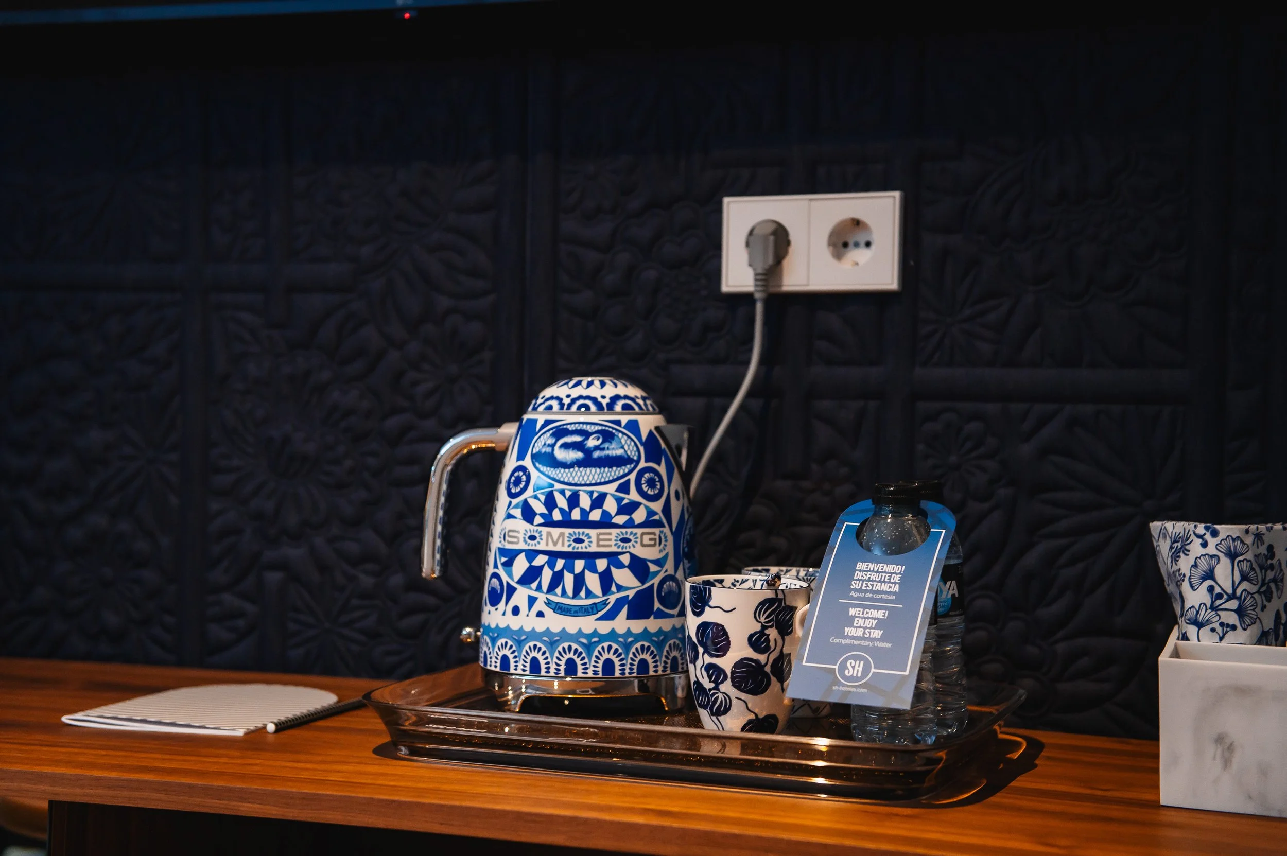 Botella de agua, taza y refri en una bandeja sobre una mesa de madera en un fondo oscuro con enchufe eléctrico.