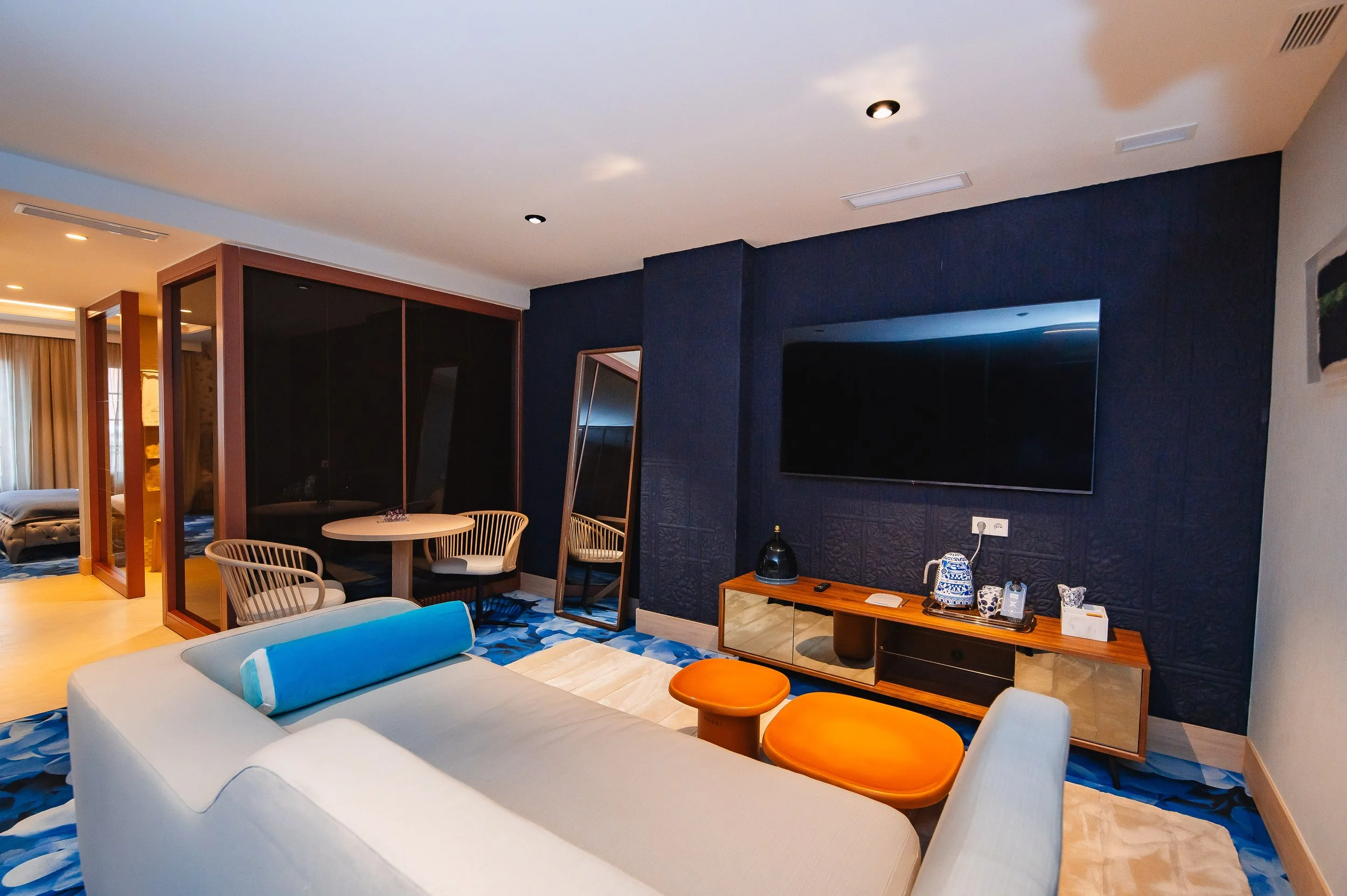 Sala de estar con sofá beige, mesa de centro color naranja, televisión grande en la pared azul, junto a un mueble de madera, en un ambiente moderno con decoración elegante.