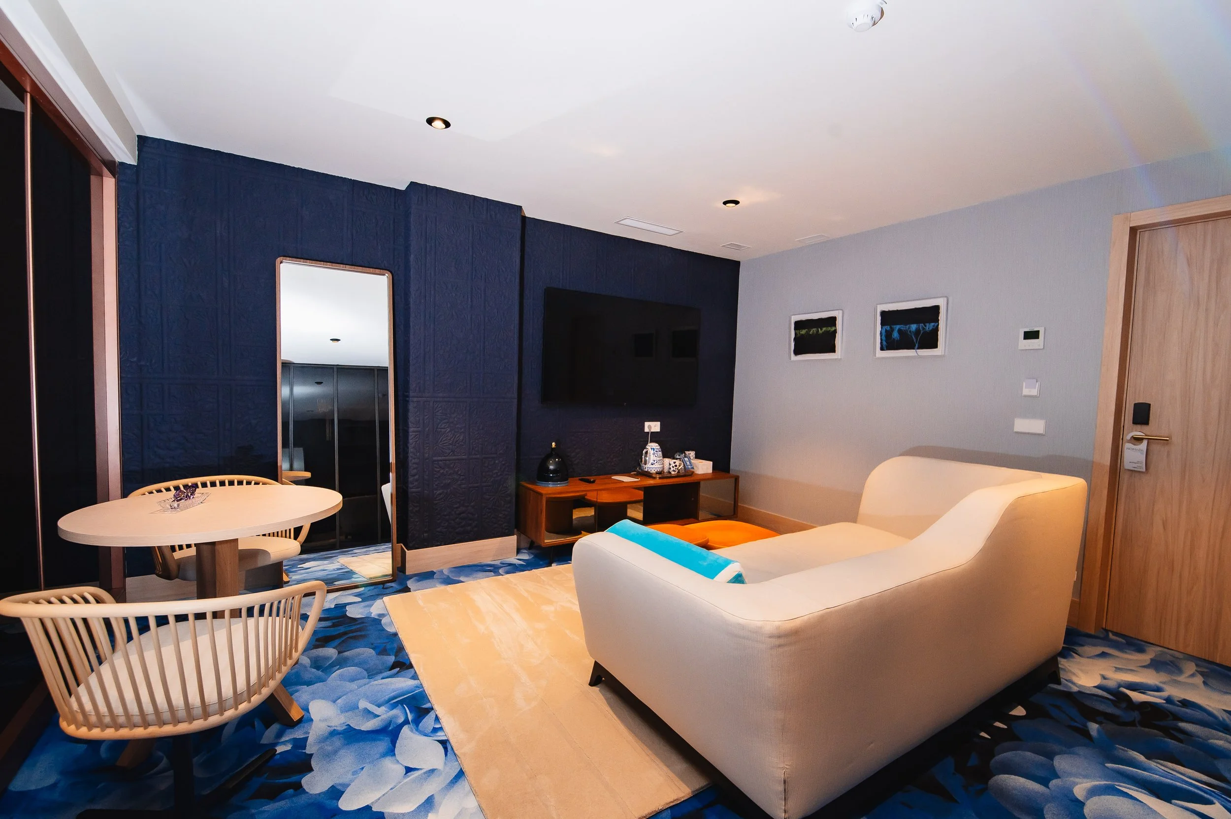 Sala de hotel con sofá beige, televisión en pared, mesa de centro, dos cuadros decorativos en la pared, y un espejo con paredes azules.