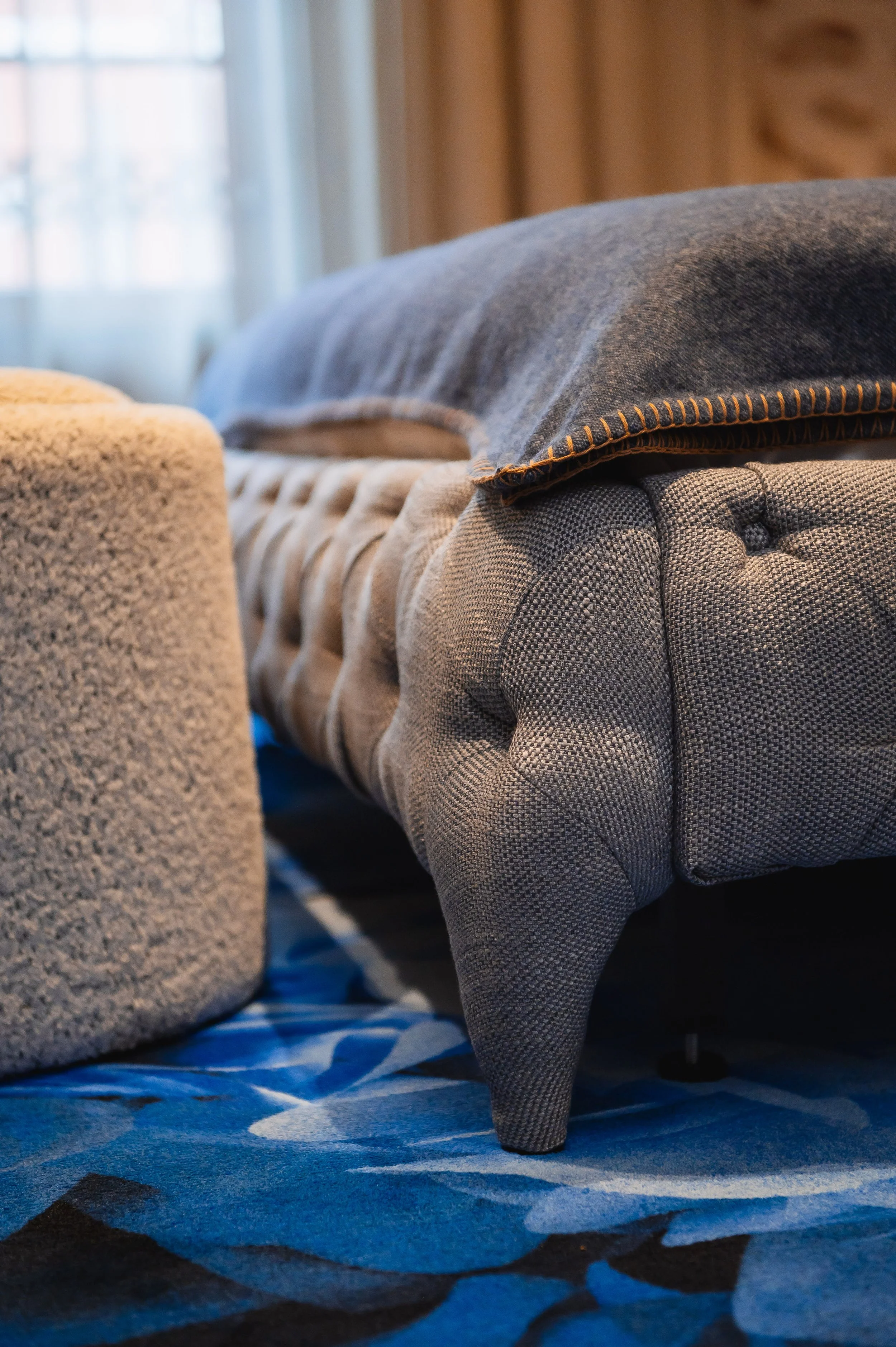 Un sofá gris de diseño clásico, con patas cortas y detalles en botones, en un salón con cortinas blancas y luz natural, sobre una alfombra con patrones en tonos azules y negros.