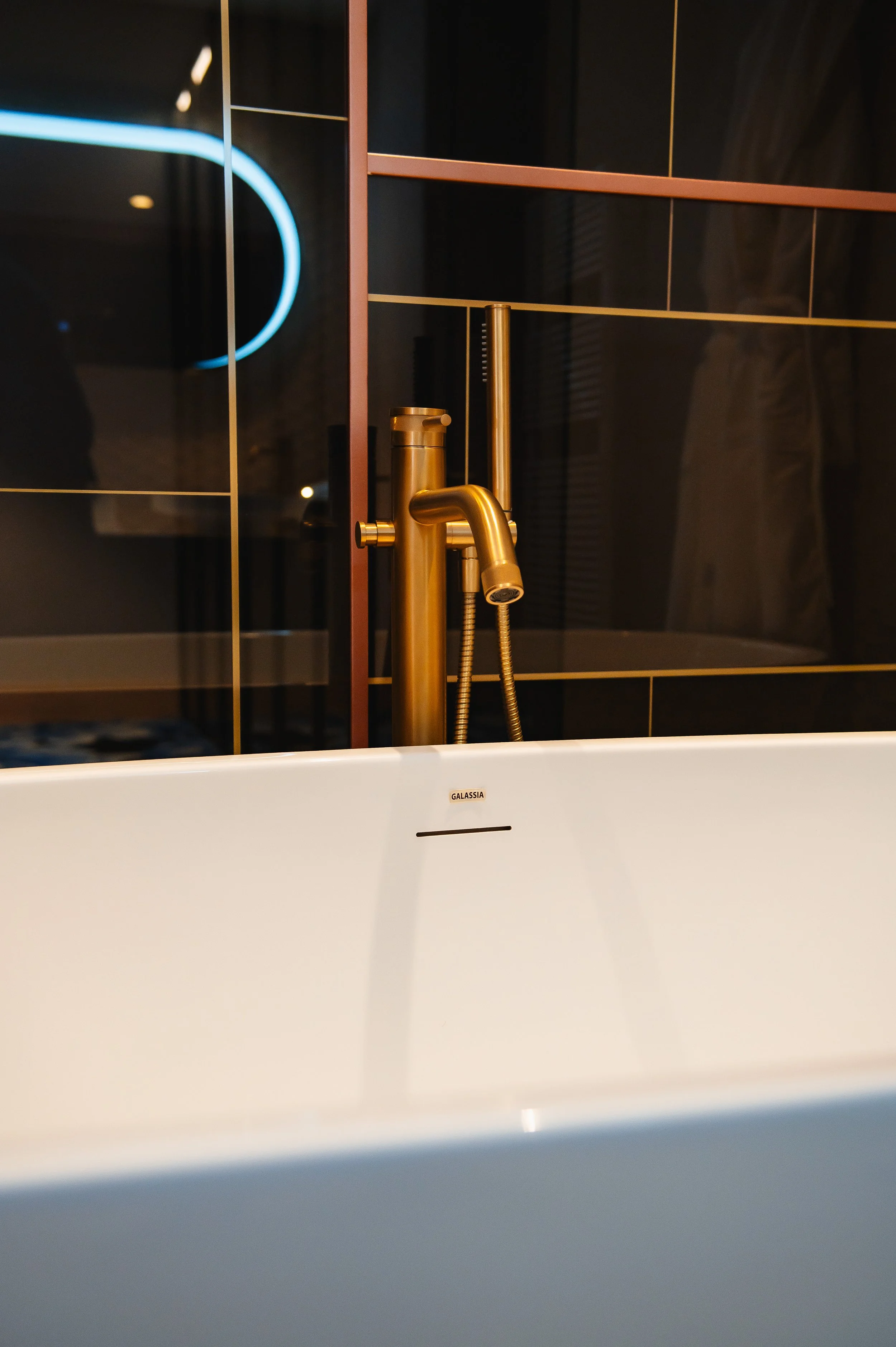 Grifo de oro en una bañera blanca, con azulejos oscuros y reflejo de luces en el fondo.