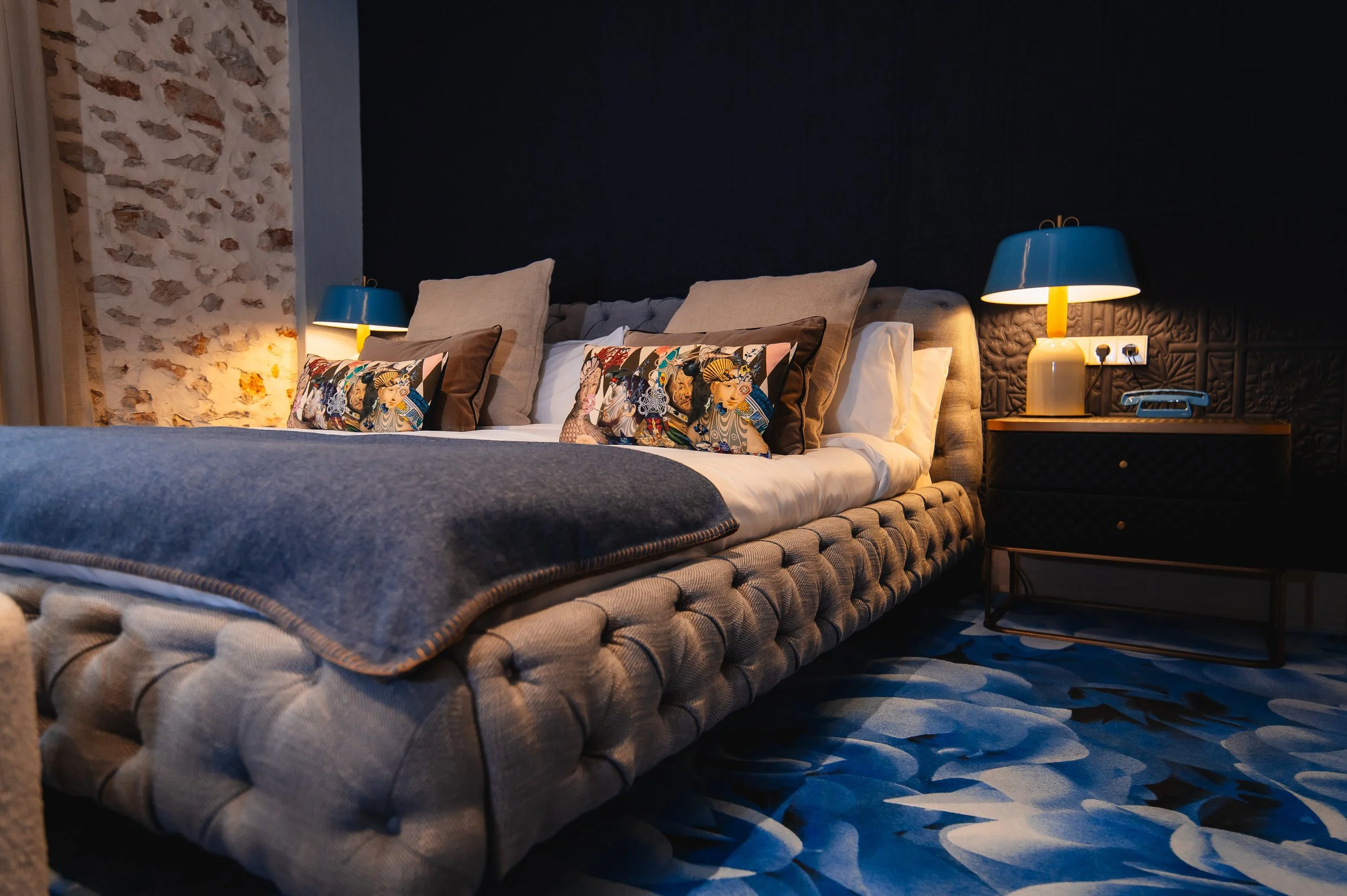 Cama de matrimonio con varios cojines decorativos, lámparas de noche, y una pared de ladrillos en un habitación moderna y elegante.