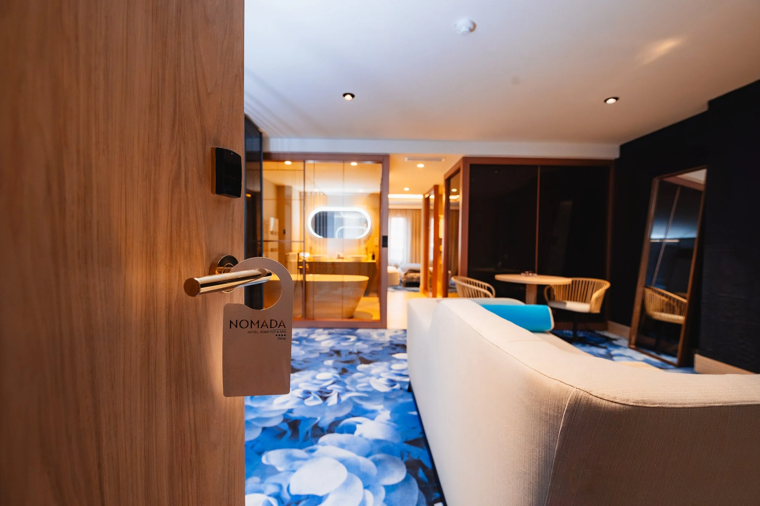 Puerta de entrada a una habitación de hotel con etiqueta que dice 'NOMADA'. Interior moderno con sillón beige, alfombra con diseño de guijarros azules y muebles de madera. En el fondo, área de baño con paredes de cristal y bañera, y una sala de estar