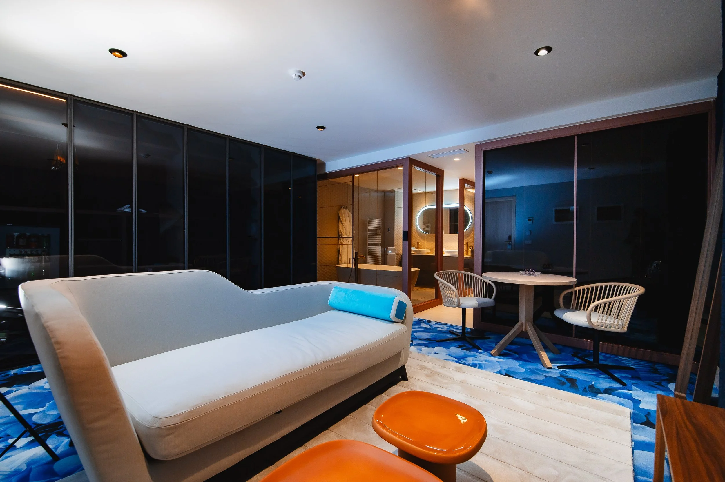 Sala moderna con sofá blanco, mesas pequeñas de madera color ámbar, sillas blancas con respaldo de varillas, alfombra azul con patrón de hojas, paredes de vidrio y un baño visible en el fondo con espejo con luz circular
