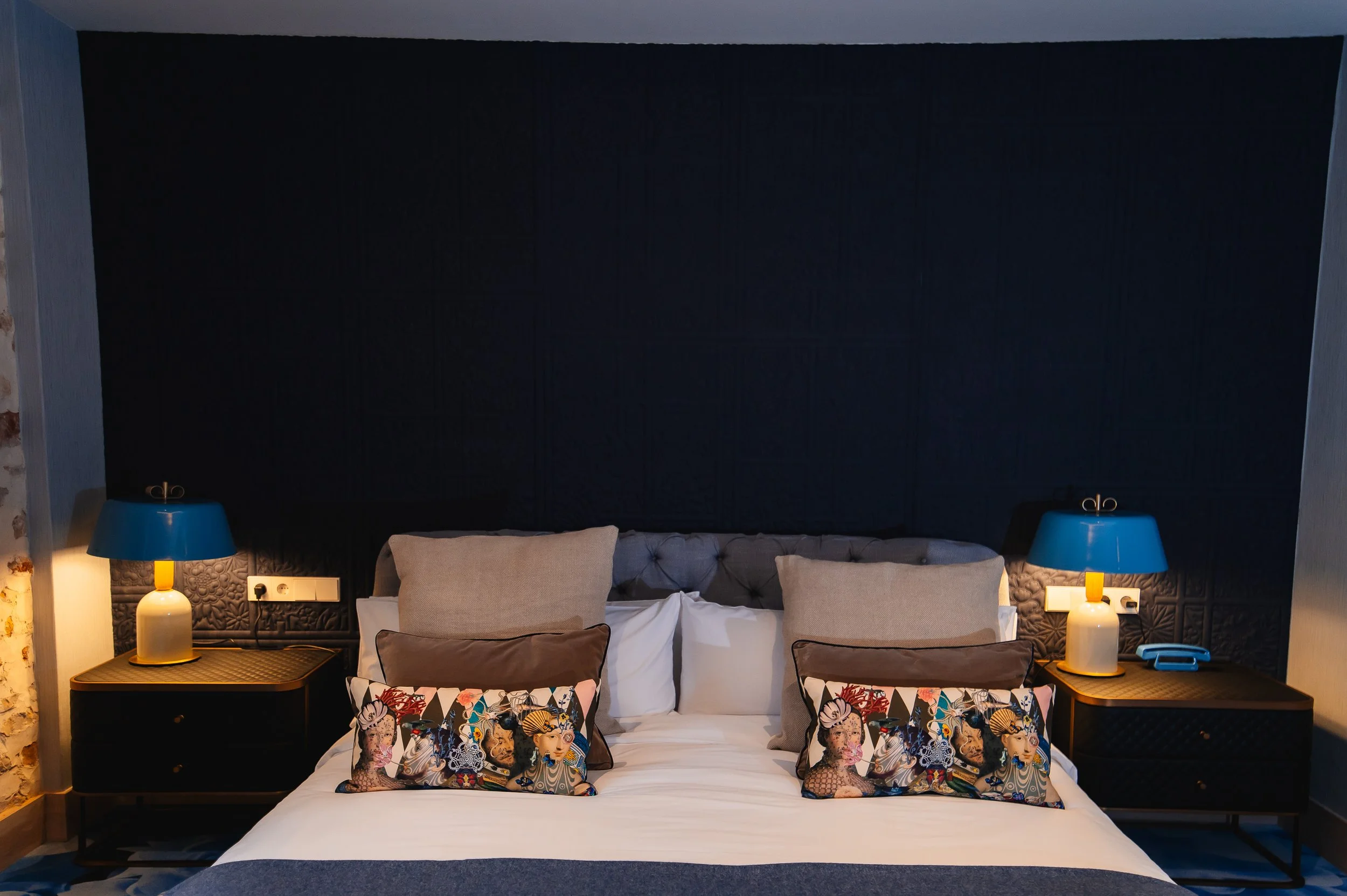 Cama con almohadas y dos lámparas azules sobre mesitas negras en un dormitorio con paredes oscuras y luz suave.