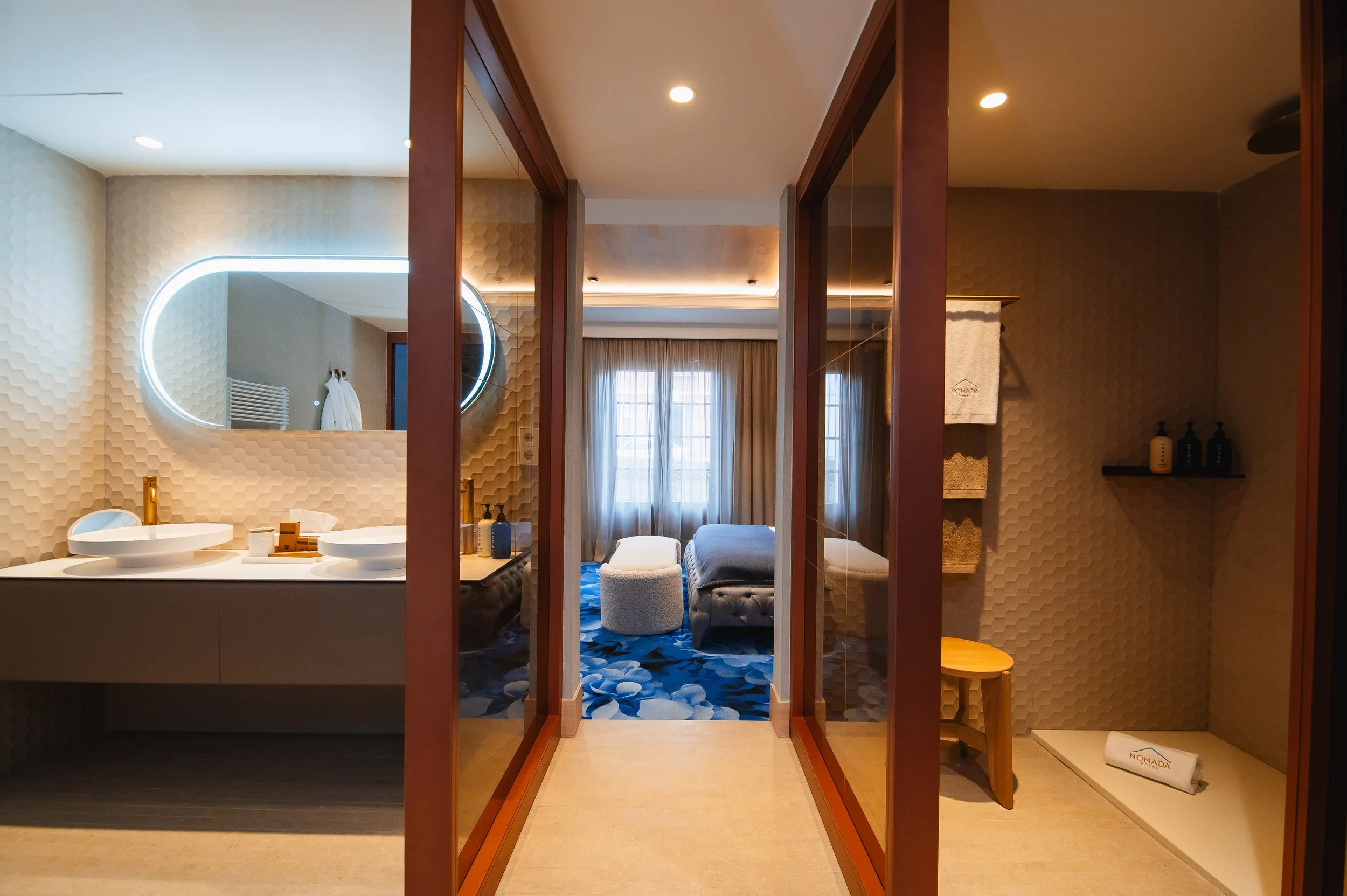 Interior de una habitación de hotel con área de baño y dormitorio separados por paredes de vidrio. El baño tiene un espejo de diseño moderno, lavabos con pedestales dorados y accesorios en tonos dorados y azules. La habitación cuenta con una cama, un