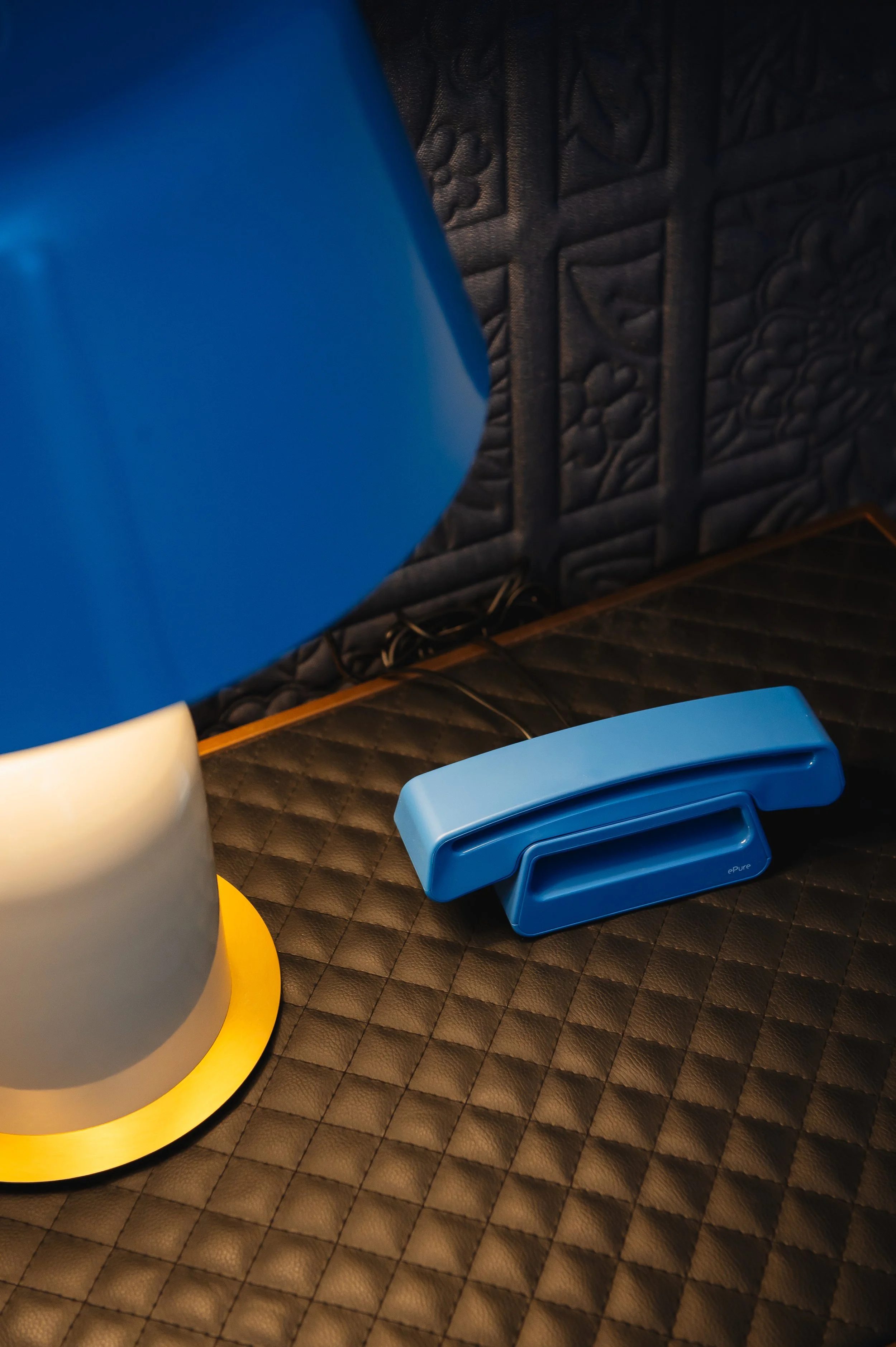 Un teléfono azul clásico de línea fija sobre una mesa con una lámpara azul y una superficie acolchada negra.